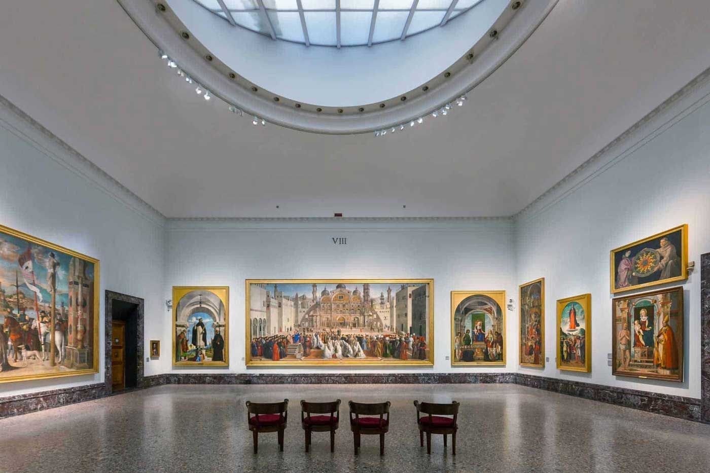 Ampia galleria museale con quadri incorniciati su pareti bianche e tre sedie sotto un lucernario.