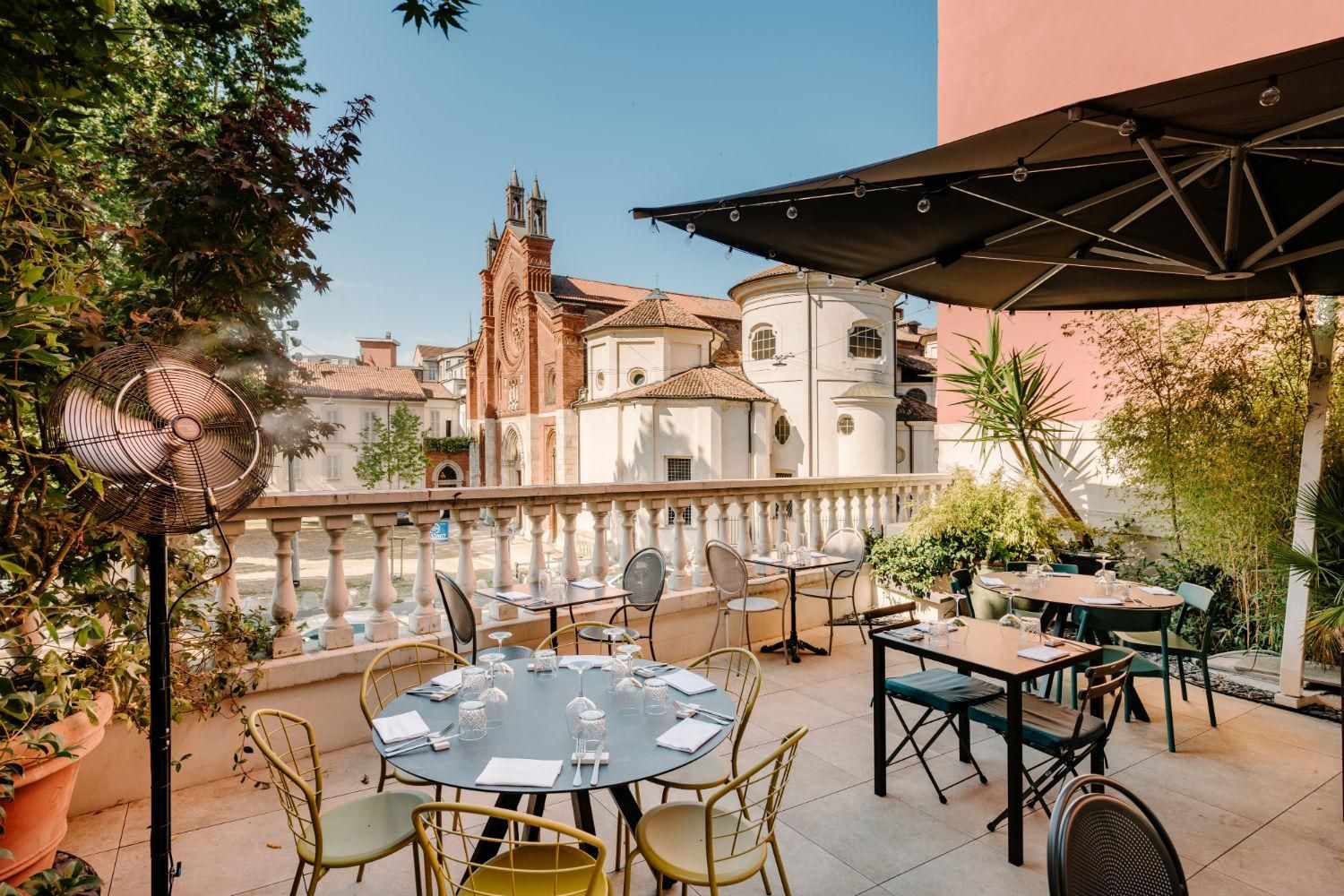 Caffè con terrazza, tavoli e sedie, affacciato su una storica piazza europea sotto un cielo azzurro.