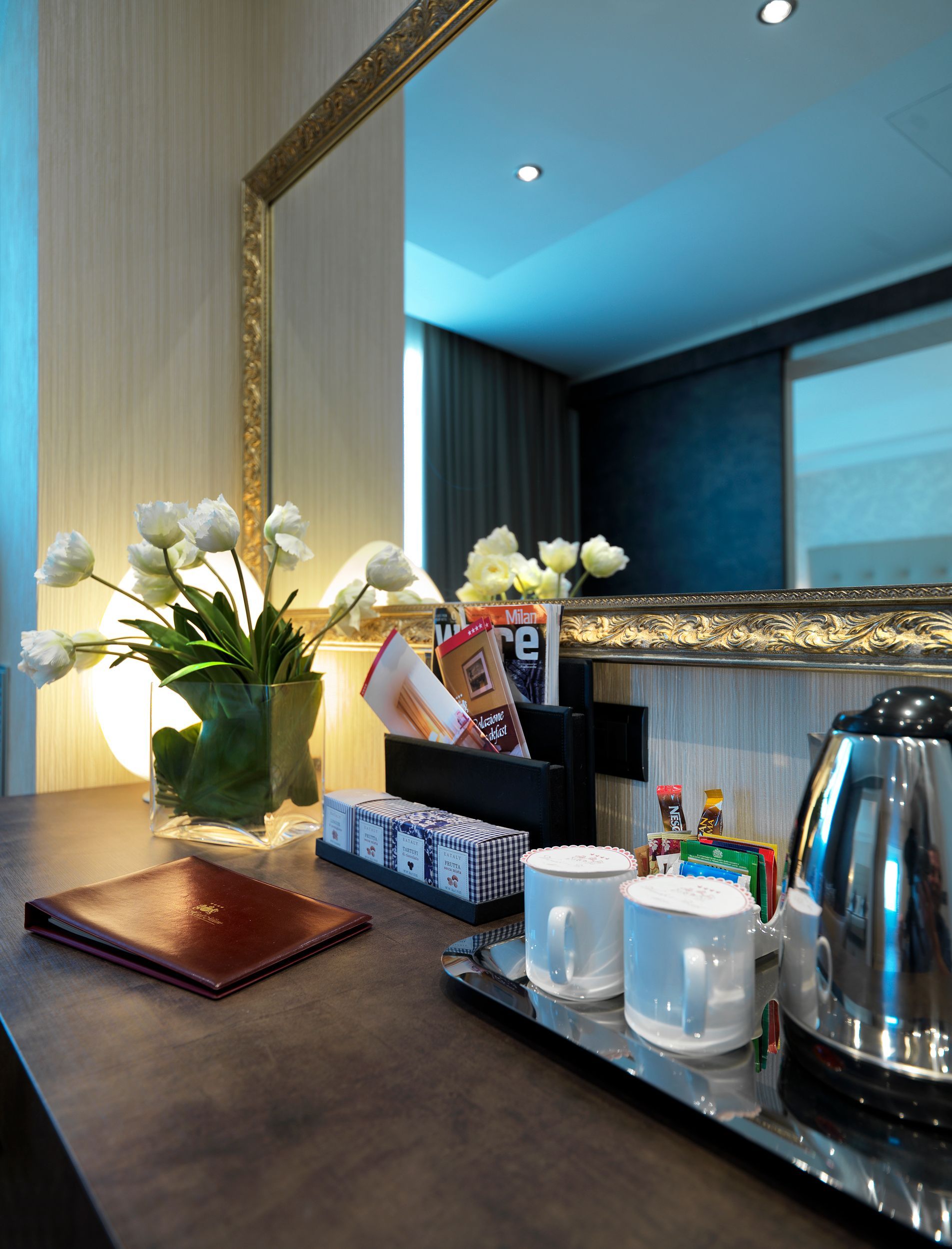 Bancone della camera d'albergo con fiori, tazze, bollitore e snack accanto a un grande specchio