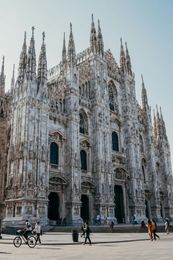 La facciata gotica bianca e riccamente decorata del Duomo di Milano, con persone e una bicicletta in primo piano.