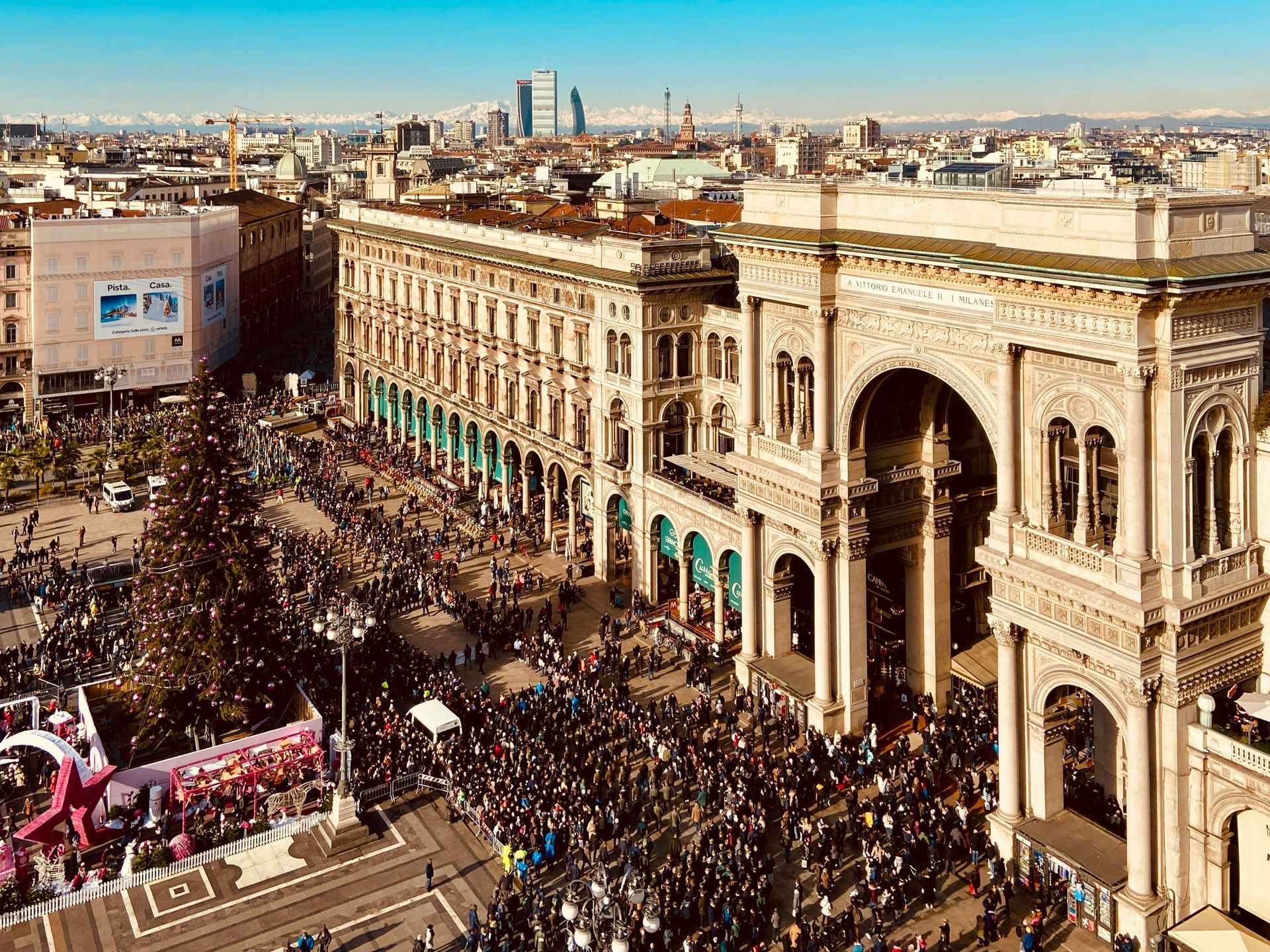 Piazza cittadina affollata accanto a un edificio storico riccamente decorato, vista dall'alto in una giornata di sole.