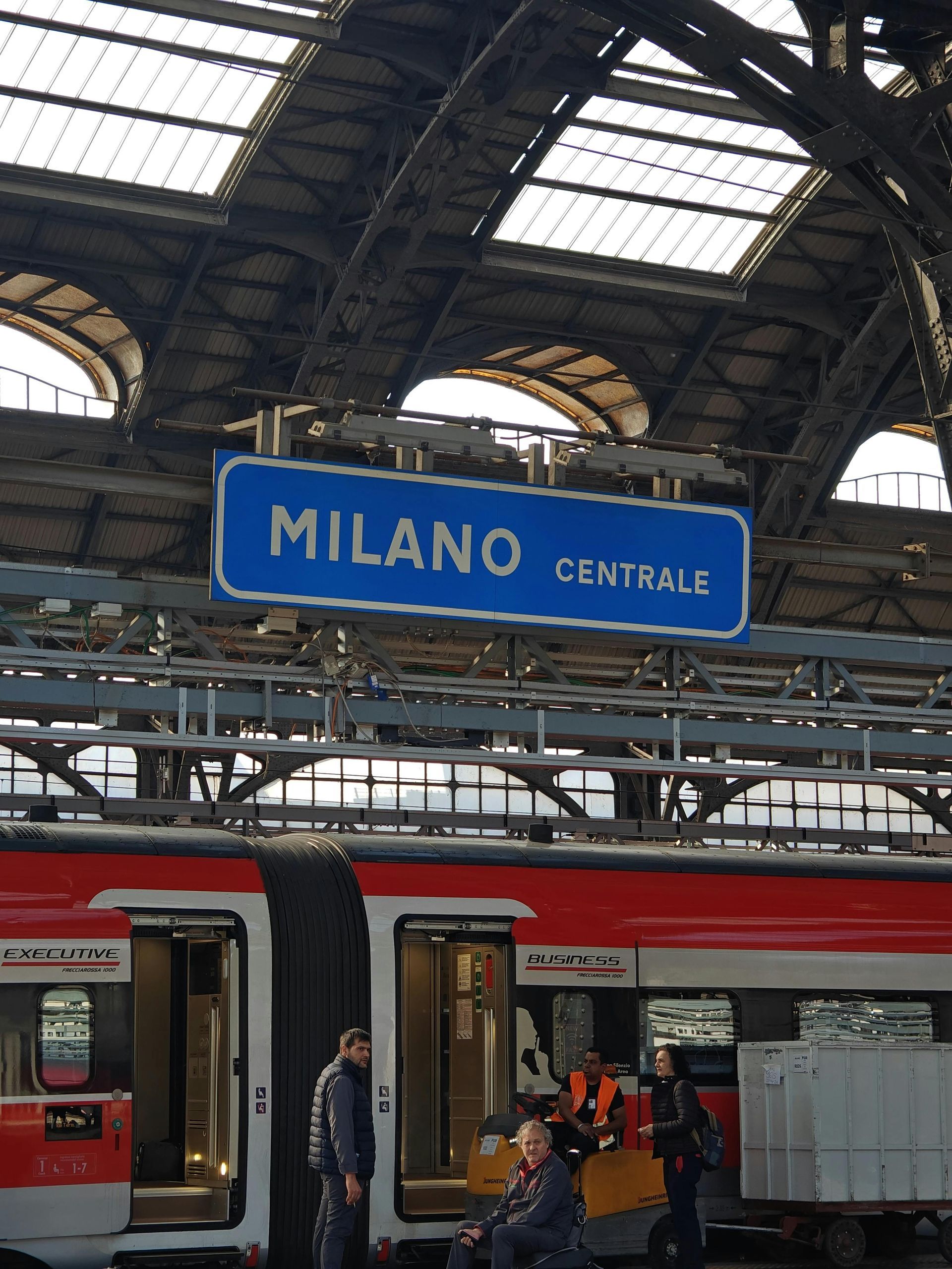 Cartello della stazione di Milano Centrale sopra un treno rosso e passeggeri sul binario.