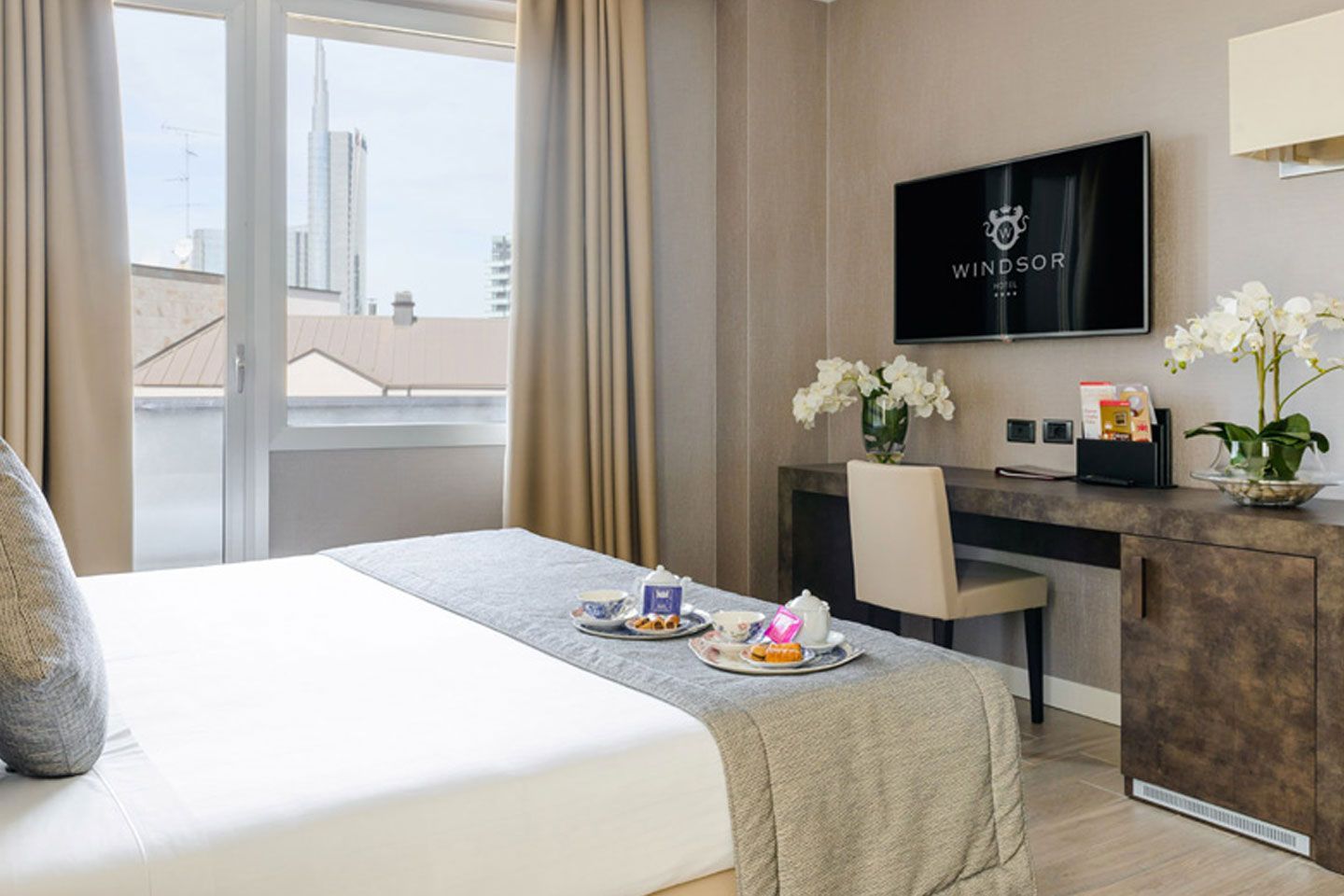 Camera d'albergo moderna con due letti singoli, una finestra e una cassettiera con TV e fiori.