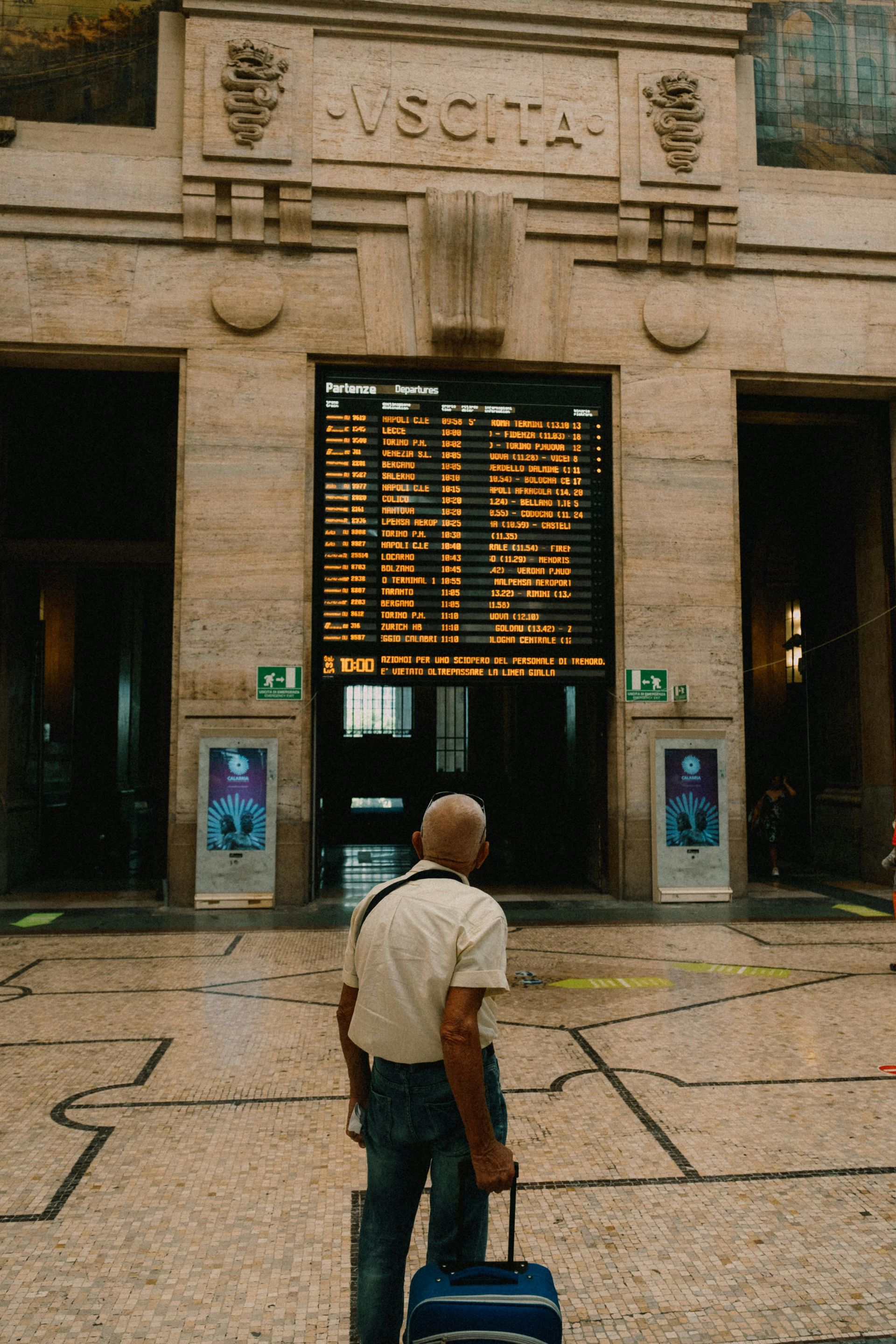 Viajero con maleta azul de pie frente a un panel luminoso de salidas de trenes en la entrada de una estación.