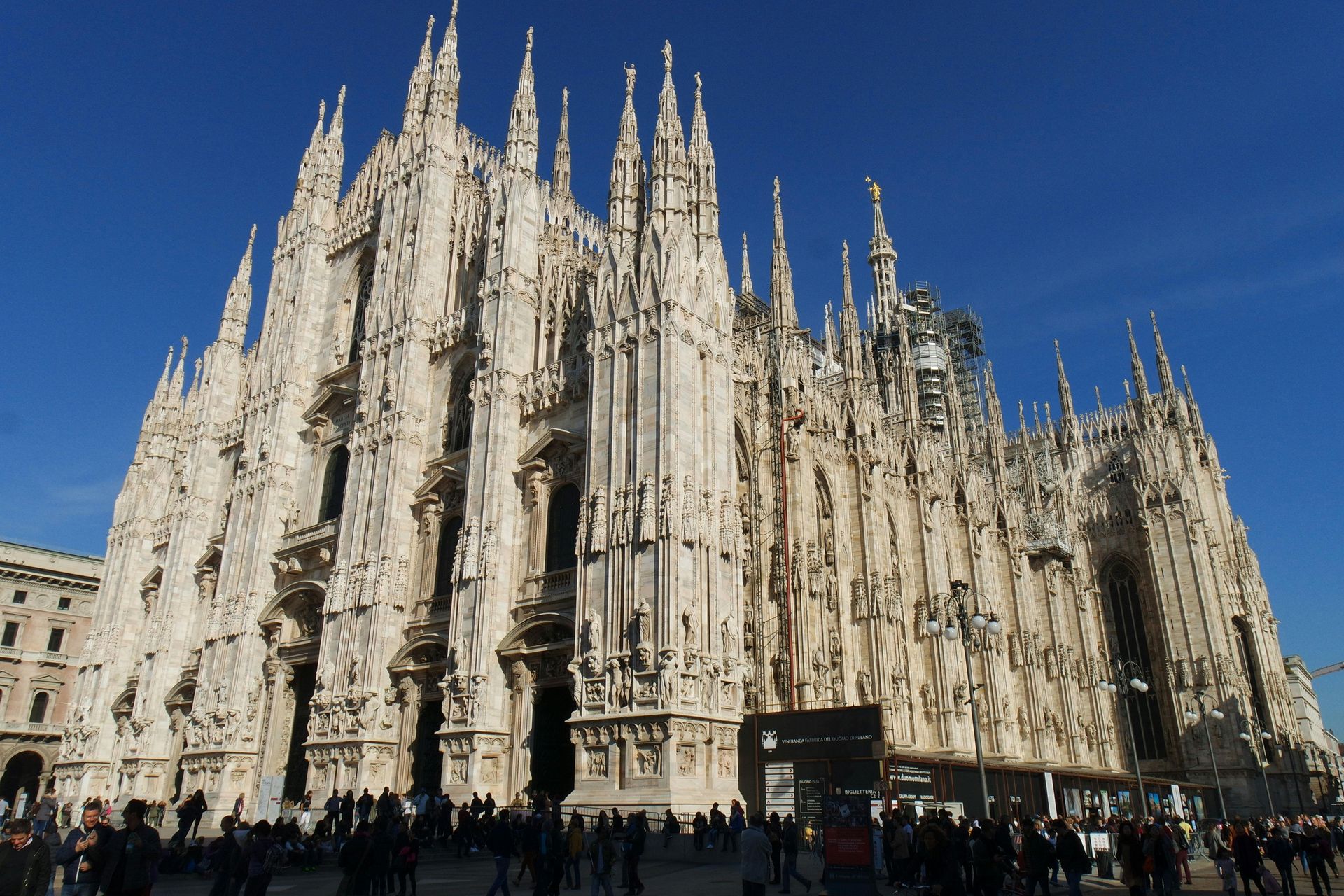 La ornamentada fachada gótica blanca de la catedral de Milán bajo un cielo azul claro, con gente reunida abajo.