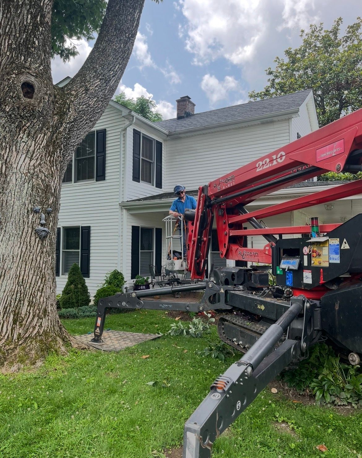 Workers using a red boom lift to reach a white house beside a large tree.