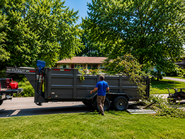 Worker by a gray dump trailer in a sunny yard with trees and a house in the background