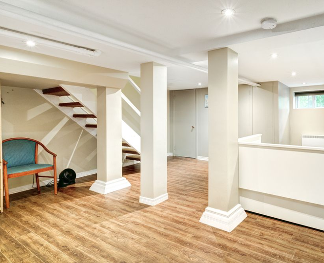 Bright finished basement with wood floors, white pillars, staircase, and a small bench under the stairs
