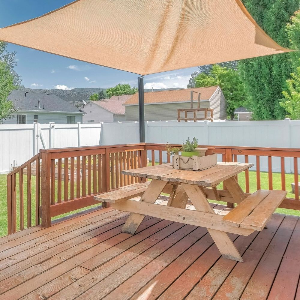 Wooden deck with picnic table under beige shade sail, surrounded by white fence and greenery.