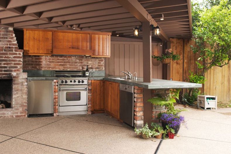 Outdoor brick patio kitchen with stainless steel grill, wooden cabinets, and a countertop under a covered roof