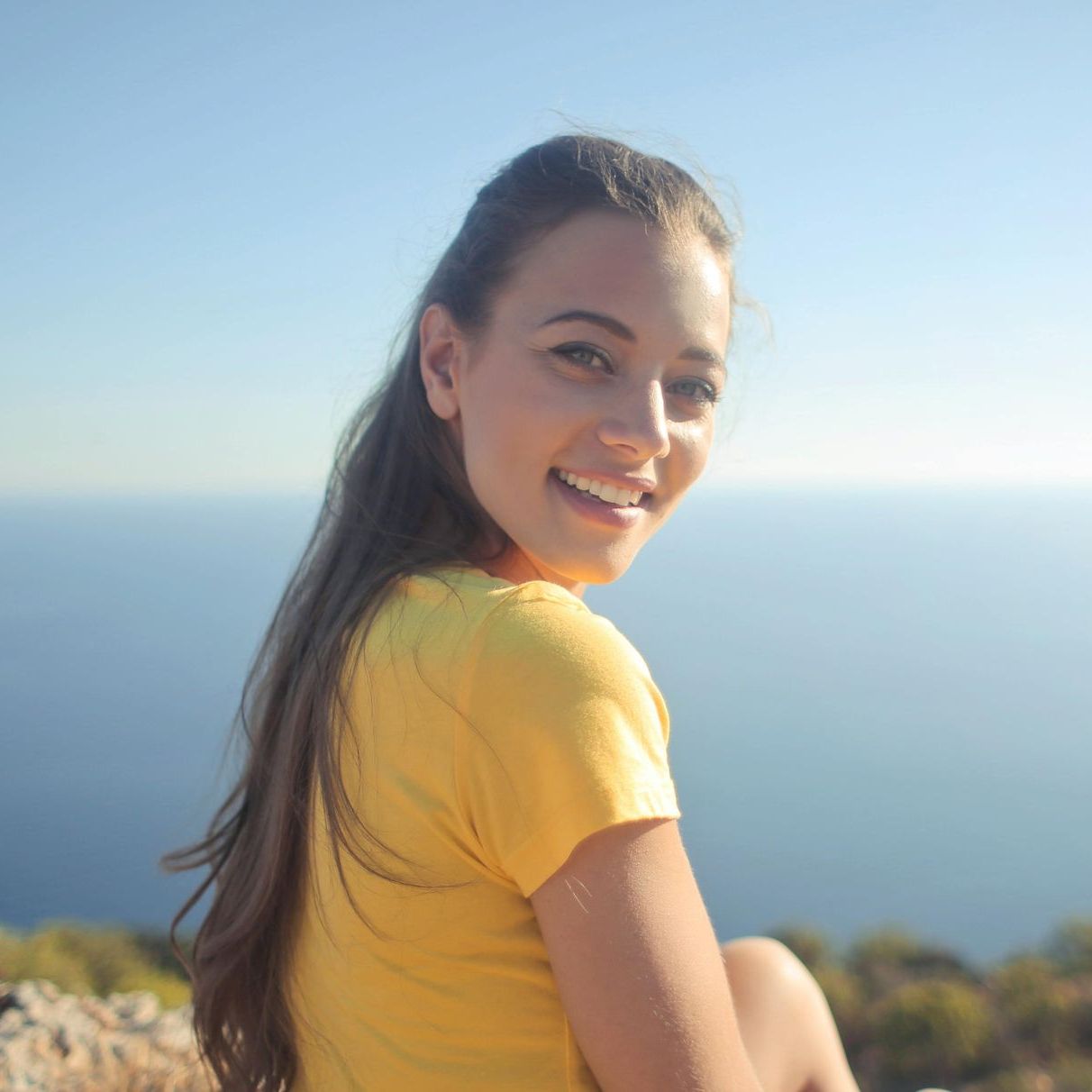 A smiling person wearing a yellow t-shirt looks back at the camera against a backdrop of the ocean.
