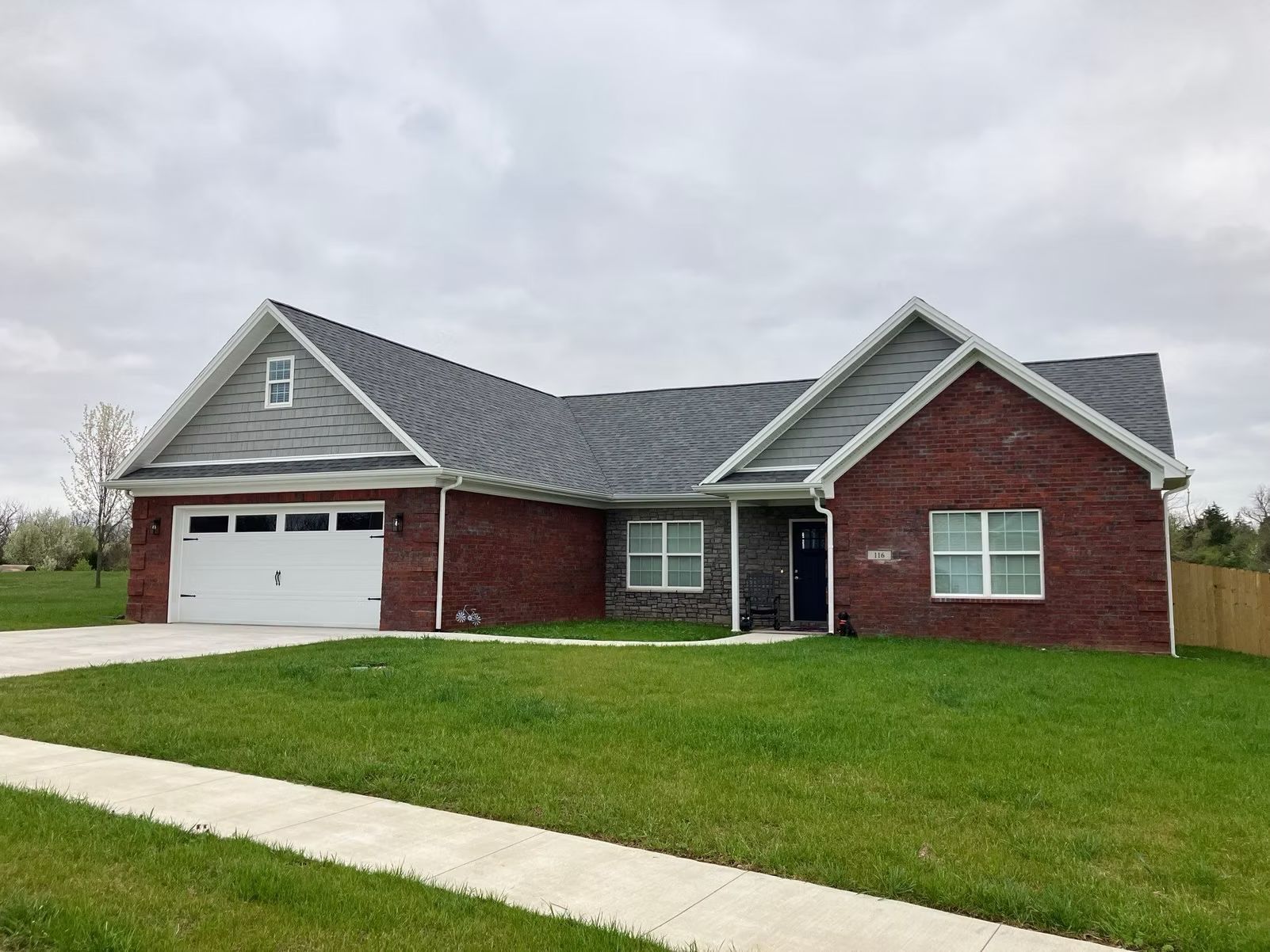 Single-story red brick house with gray roof, white garage, and green lawn under cloudy sky