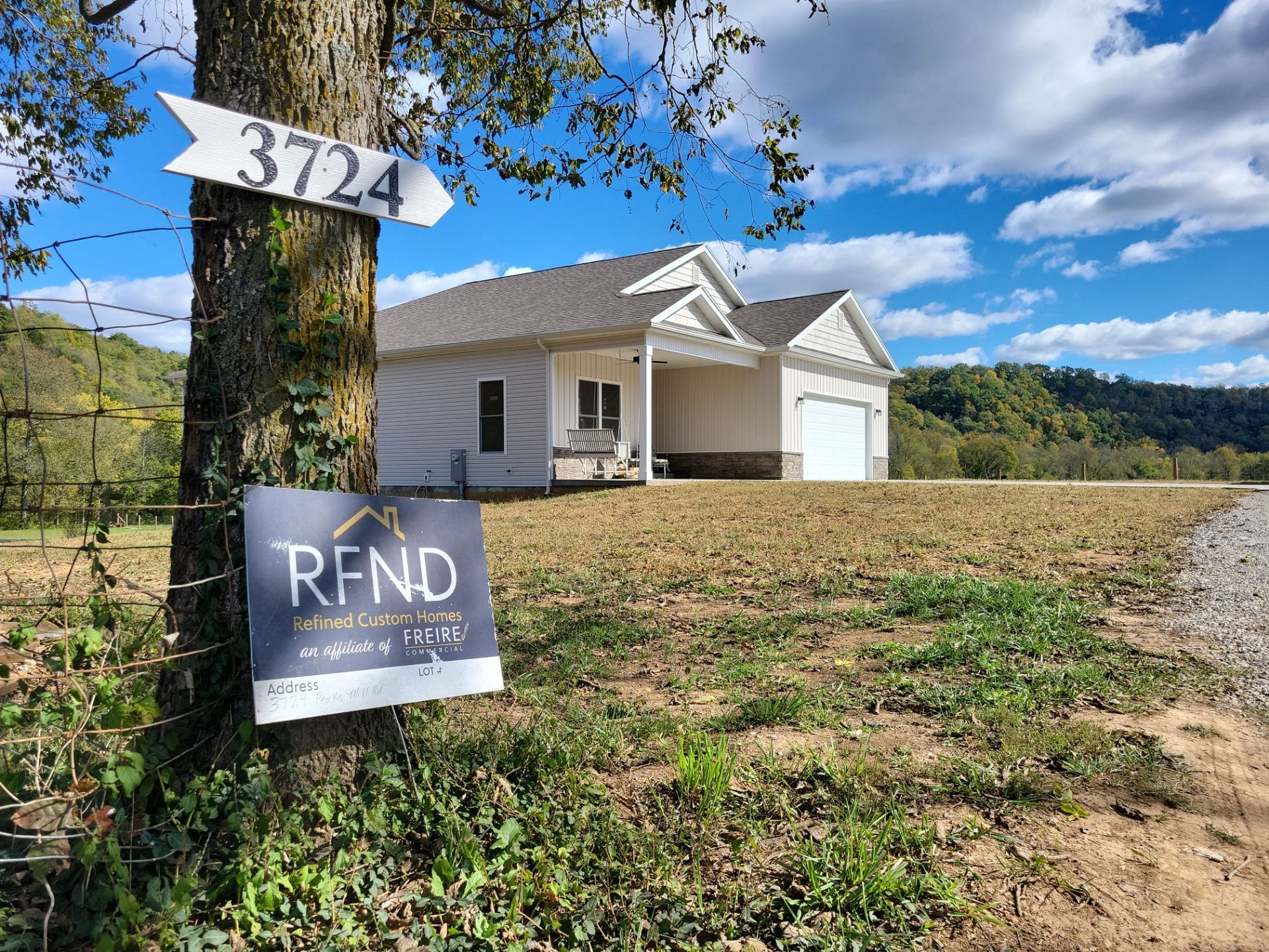 House under construction with yard signs and a tree-lined rural lot under a blue sky