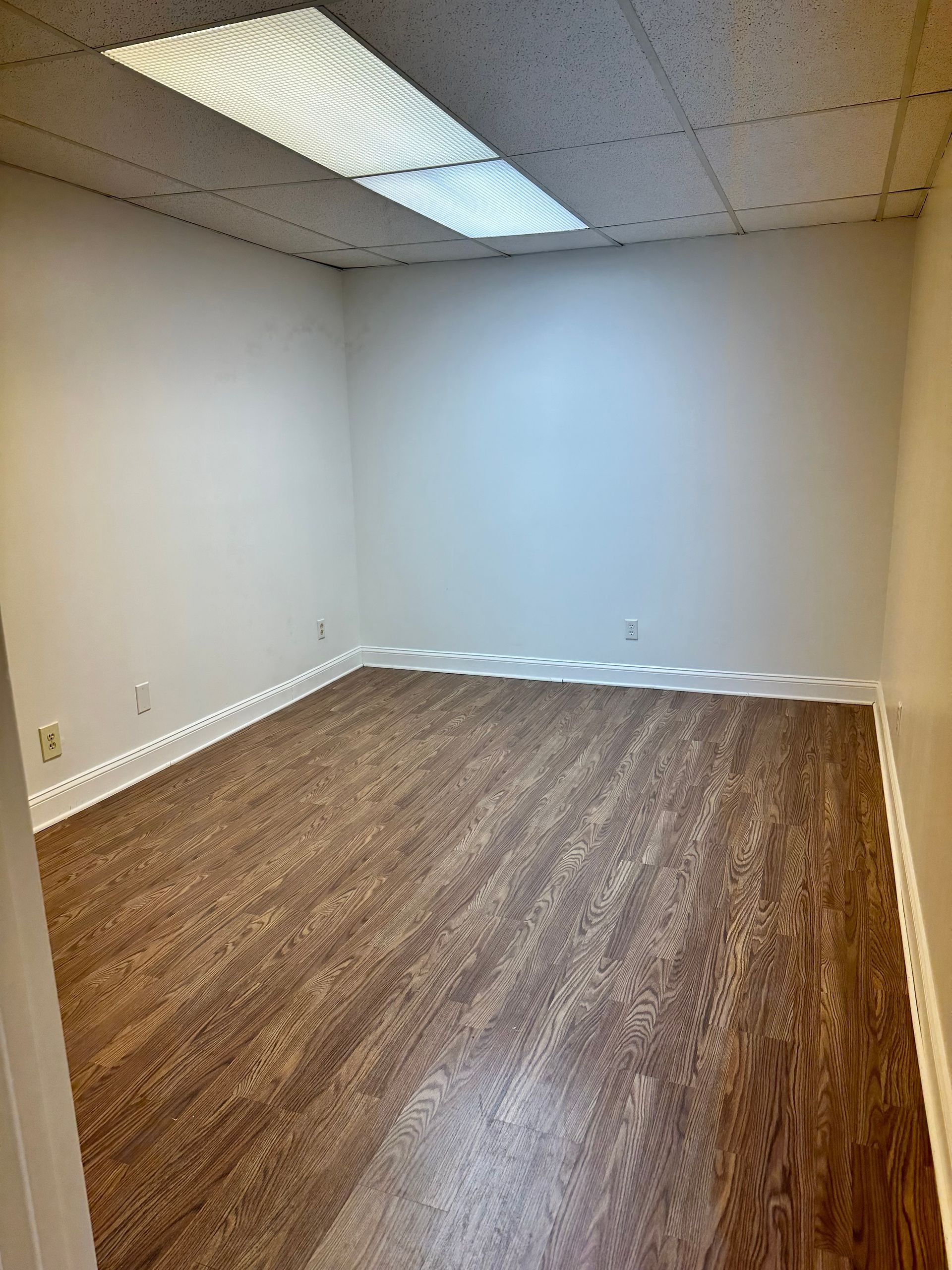 Empty office room with white walls, brown wood floor, and fluorescent ceiling light