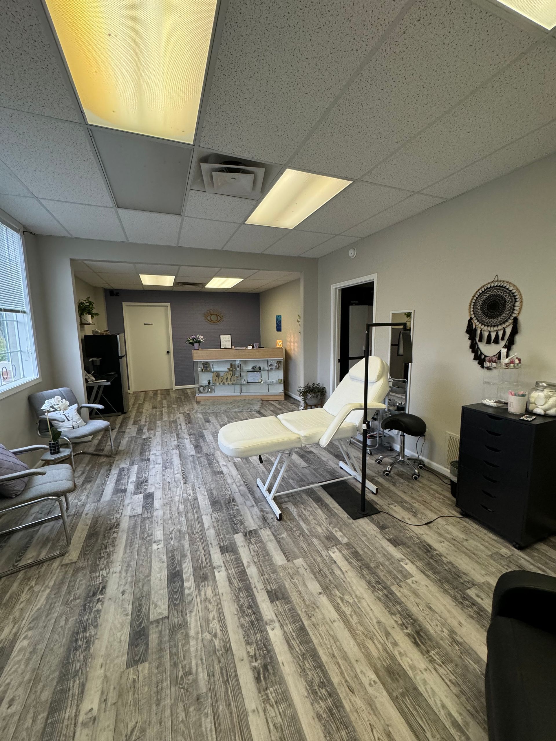 Small reception or treatment room with chairs, desk, wood-look flooring, and fluorescent ceiling lights.