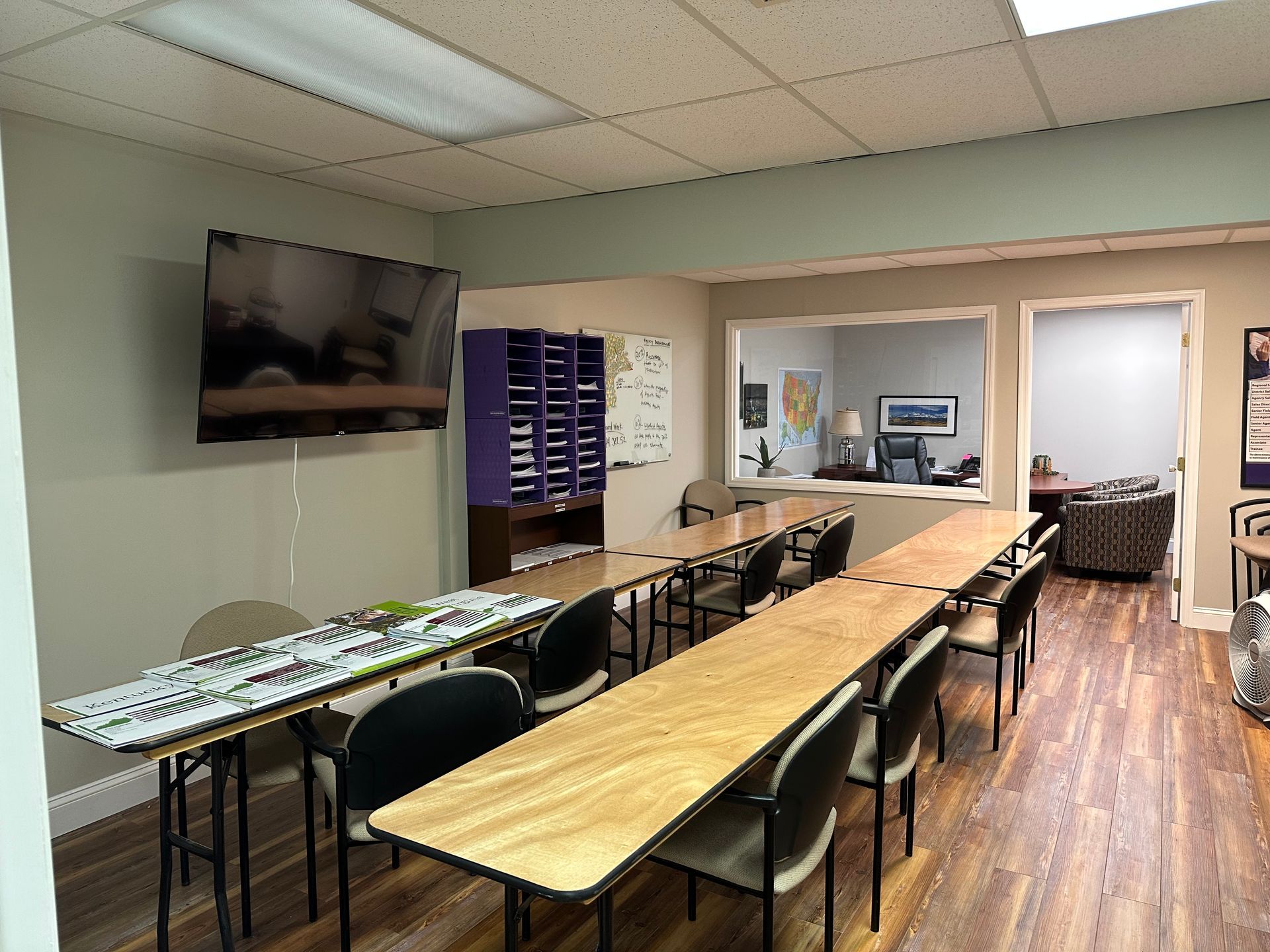Empty meeting room with long tables, chairs, wall-mounted TV, and wood flooring