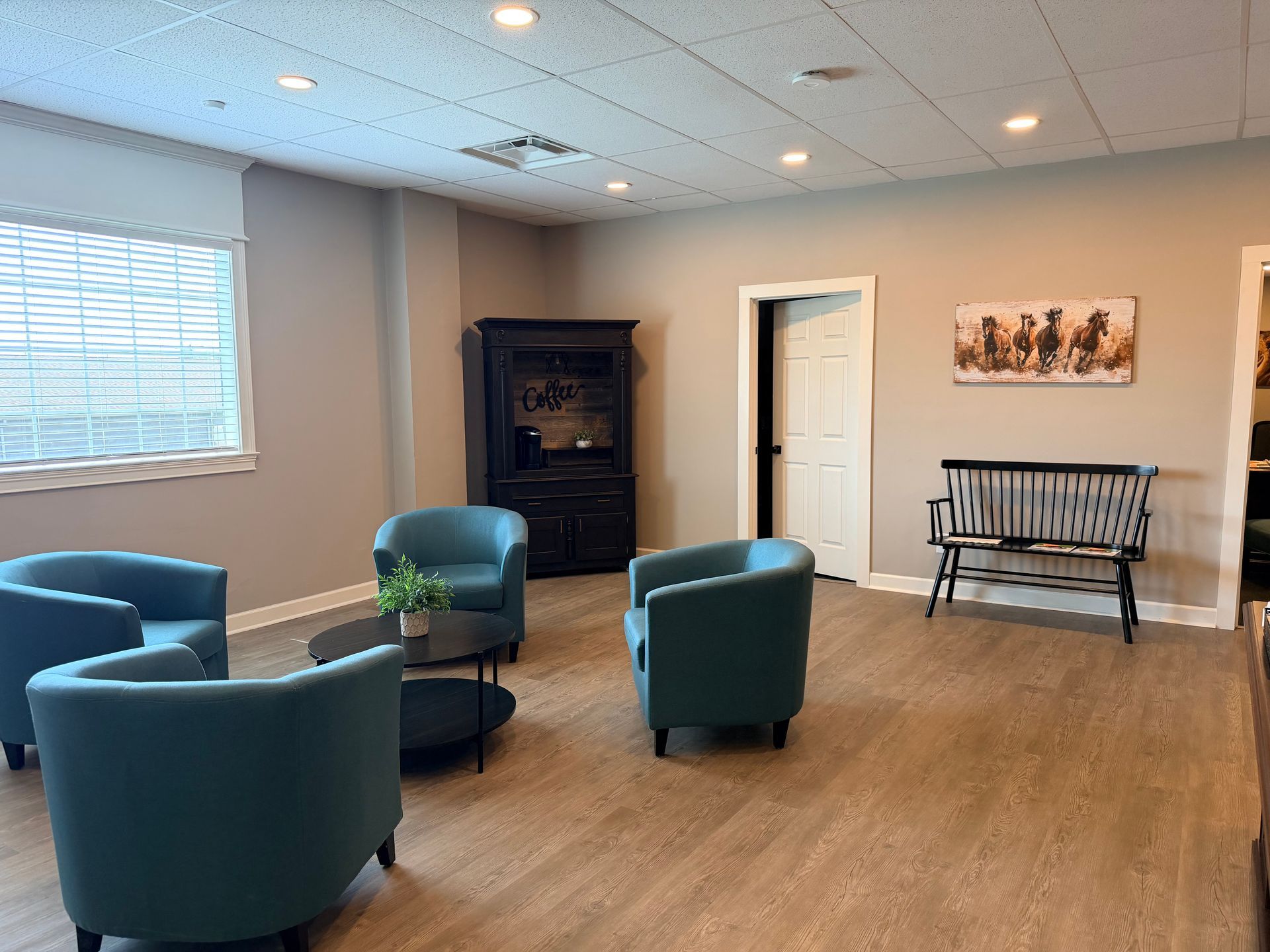 Empty waiting room with teal chairs, wood floor, window blinds, and a bench against the wall.
