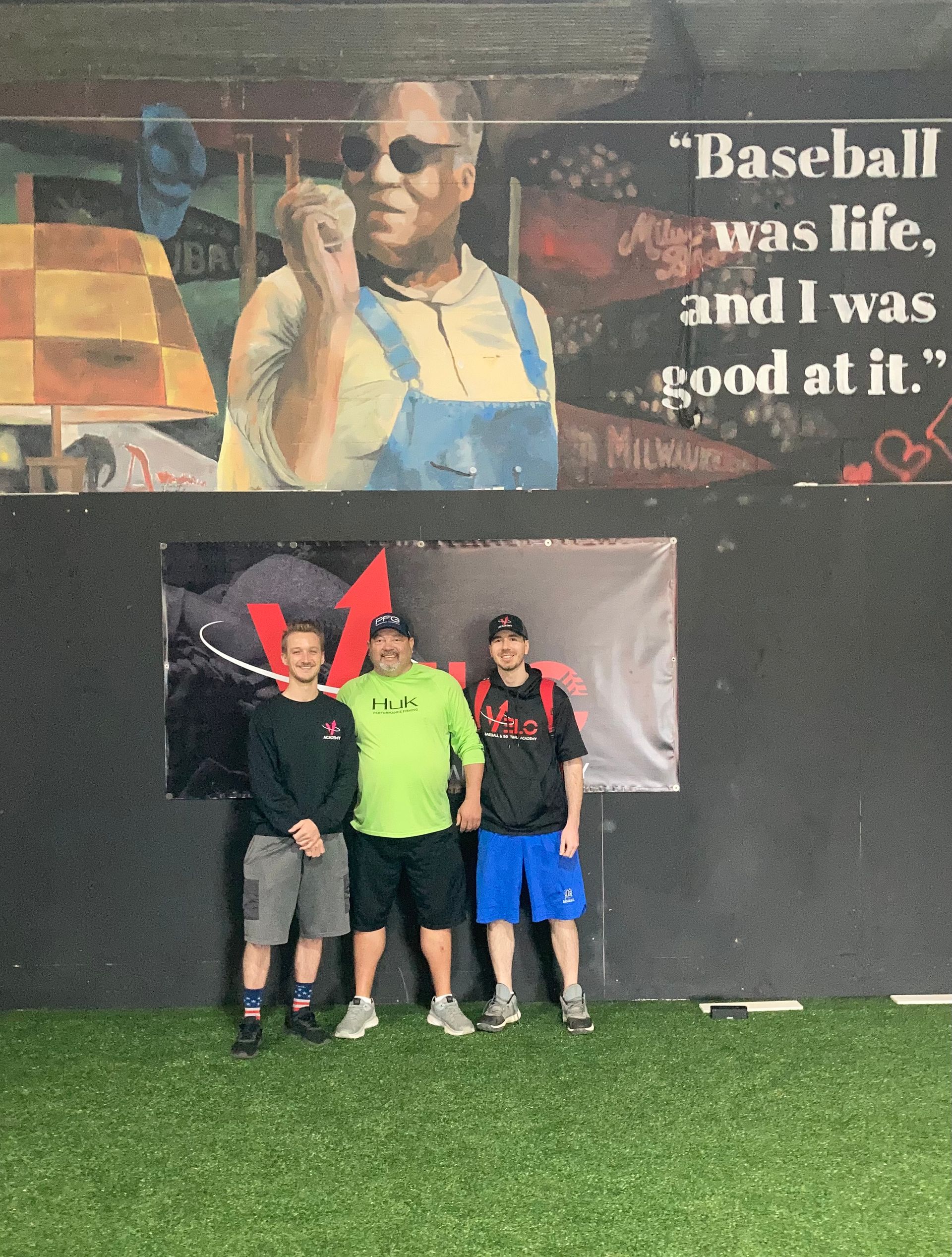Three people posing in front of a baseball mural on indoor turf, one holding a red sign.