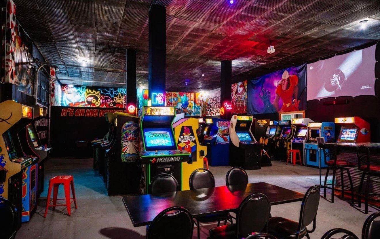 Neon-lit arcade with rows of game machines, stools, and colorful wall art in a dark room