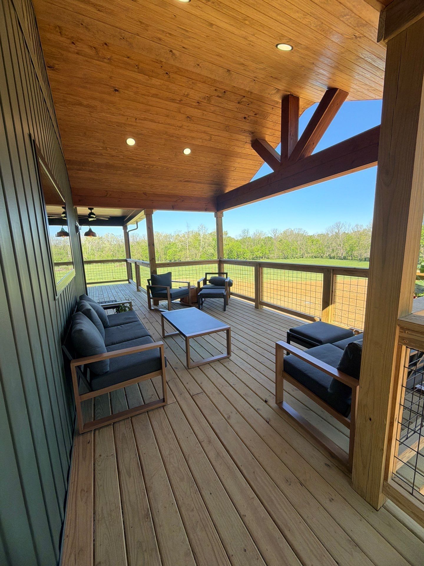 Covered wooden porch with black seating, coffee table, and scenic field view under a vaulted ceiling.