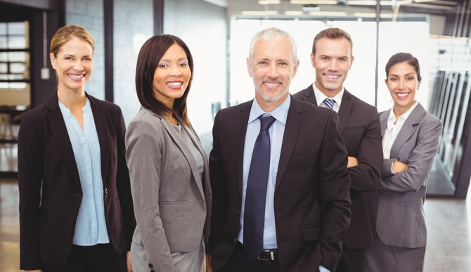 Five business professionals in suits stand smiling in a bright office lobby.