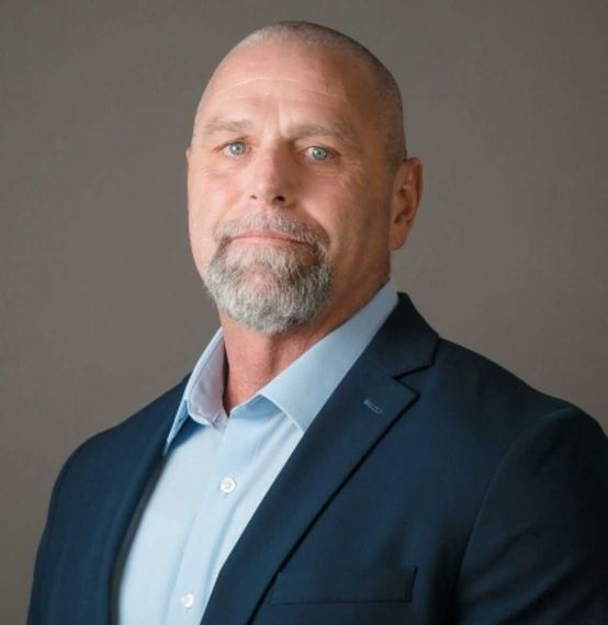 Professional headshot of a bald man in a navy suit and light blue shirt against a gray background