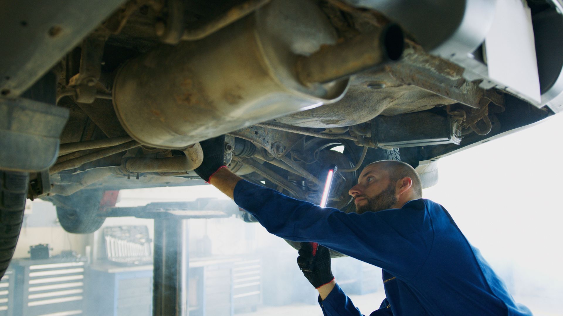 Mechanic in blue uniform inspecting undercarriage of a vehicle with a flashlight in a repair shop