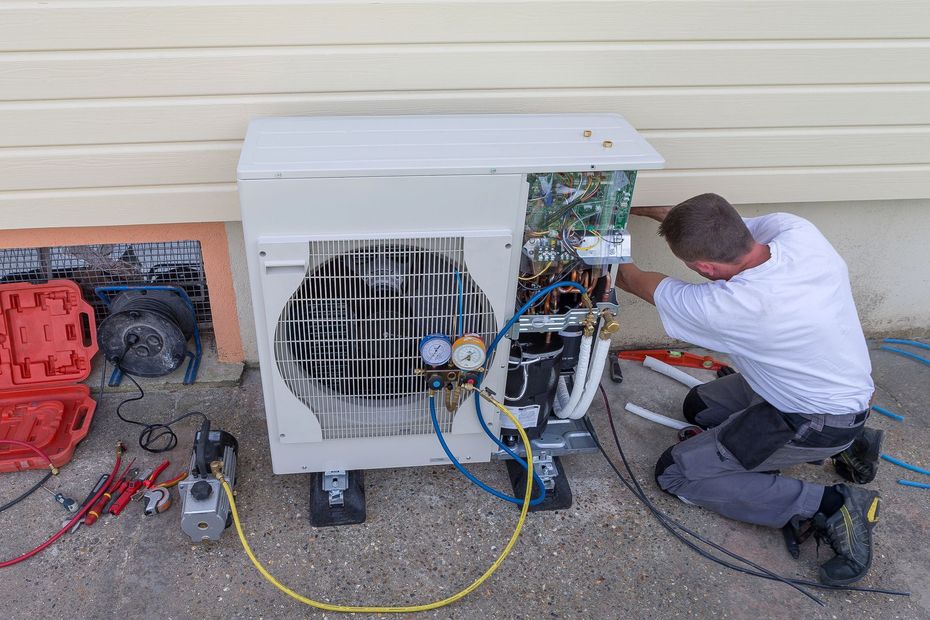 Un tecnico è inginocchiato accanto a una grande unità esterna di climatizzazione, intento a riparare i cavi scoperti utilizzando attrezzi e tubi flessibili che si trovano nelle vicinanze.