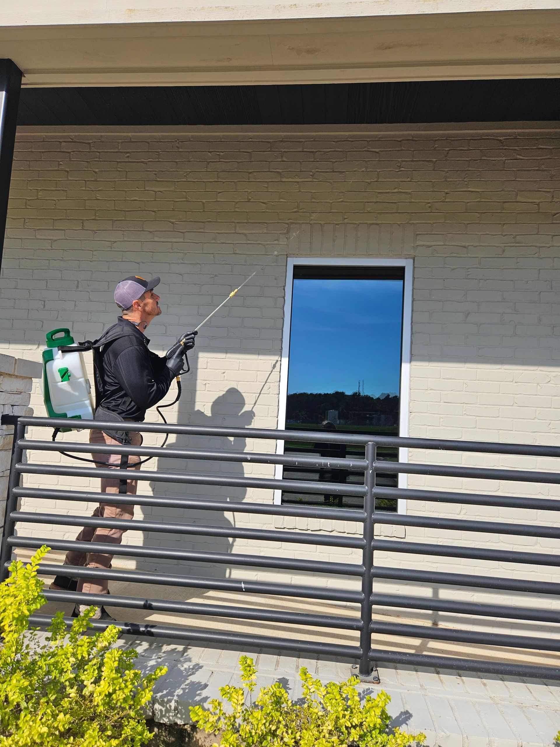 Person using a backpack sprayer to treat a beige building wall near a window and balcony railings
