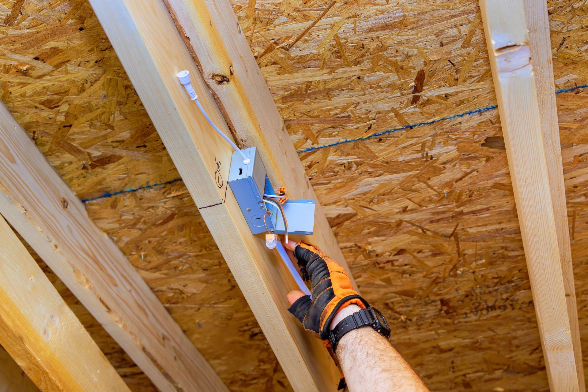 Gloved hand using a blue power tool to fasten a metal bracket to wooden ceiling joists.