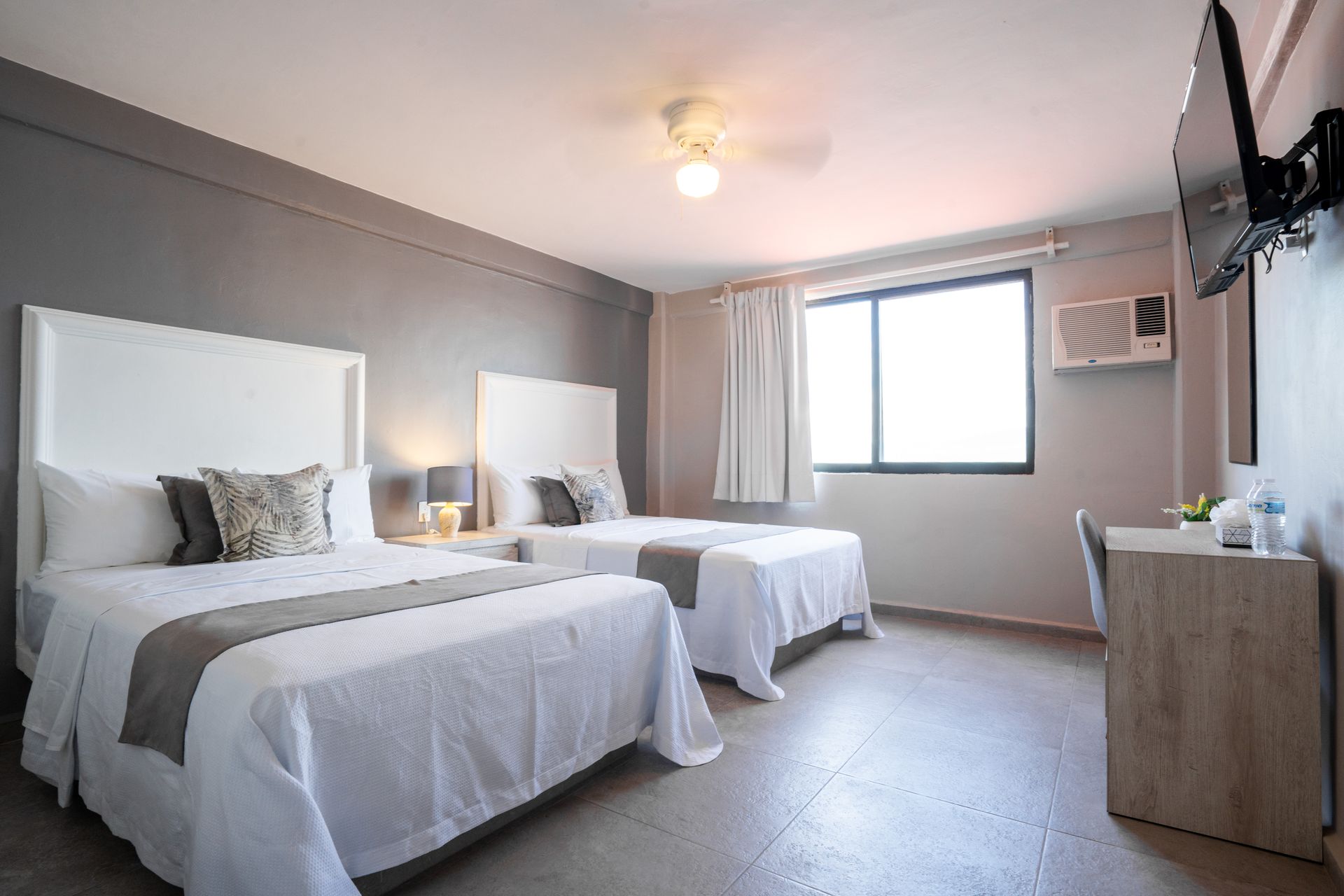 Dos camas queen en una habitación de hotel con paredes grises, escritorio, ventana y televisor montado en la pared.