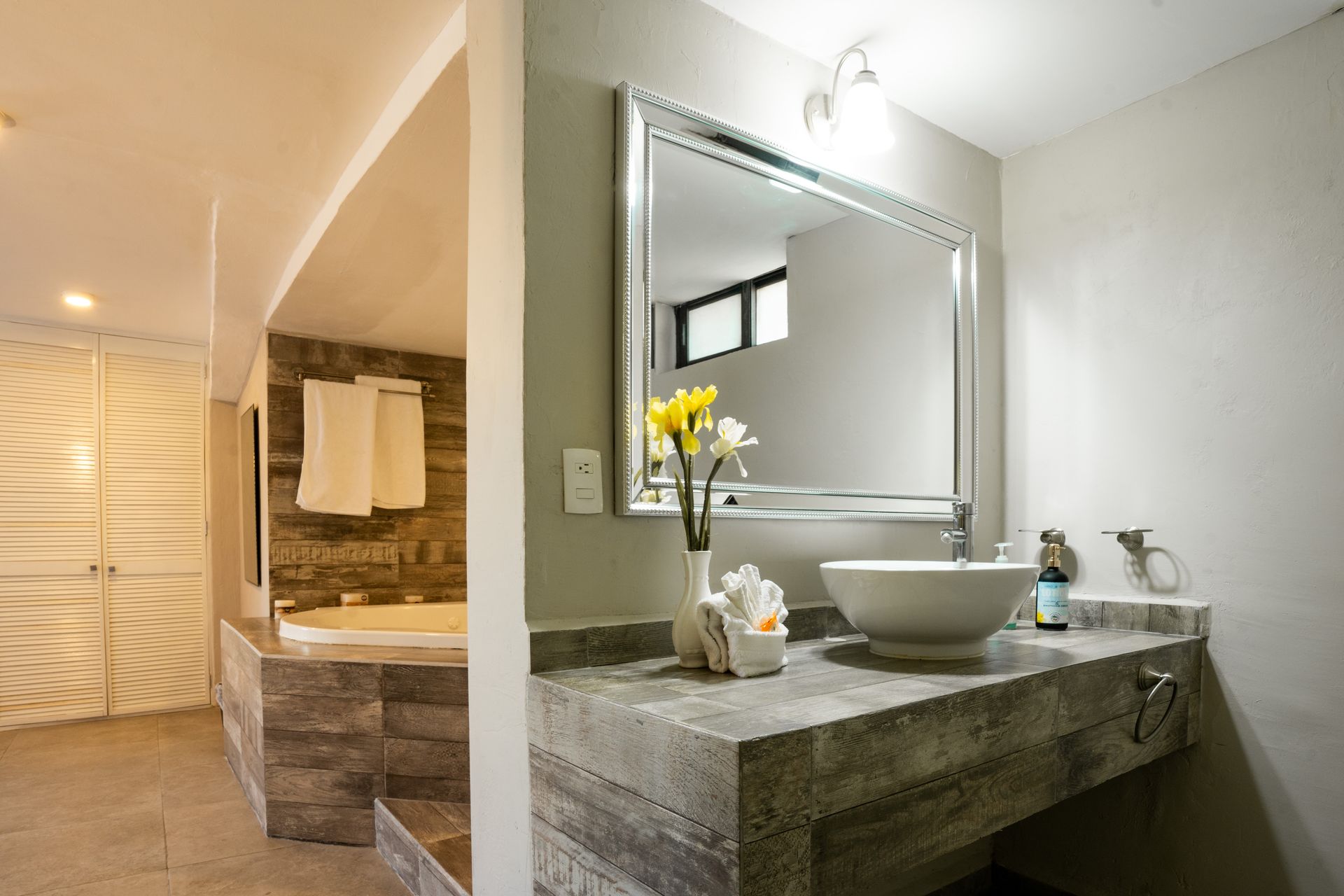 Baño con lavabo, espejo y bañera. Tonos neutros, encimera de piedra y flores.