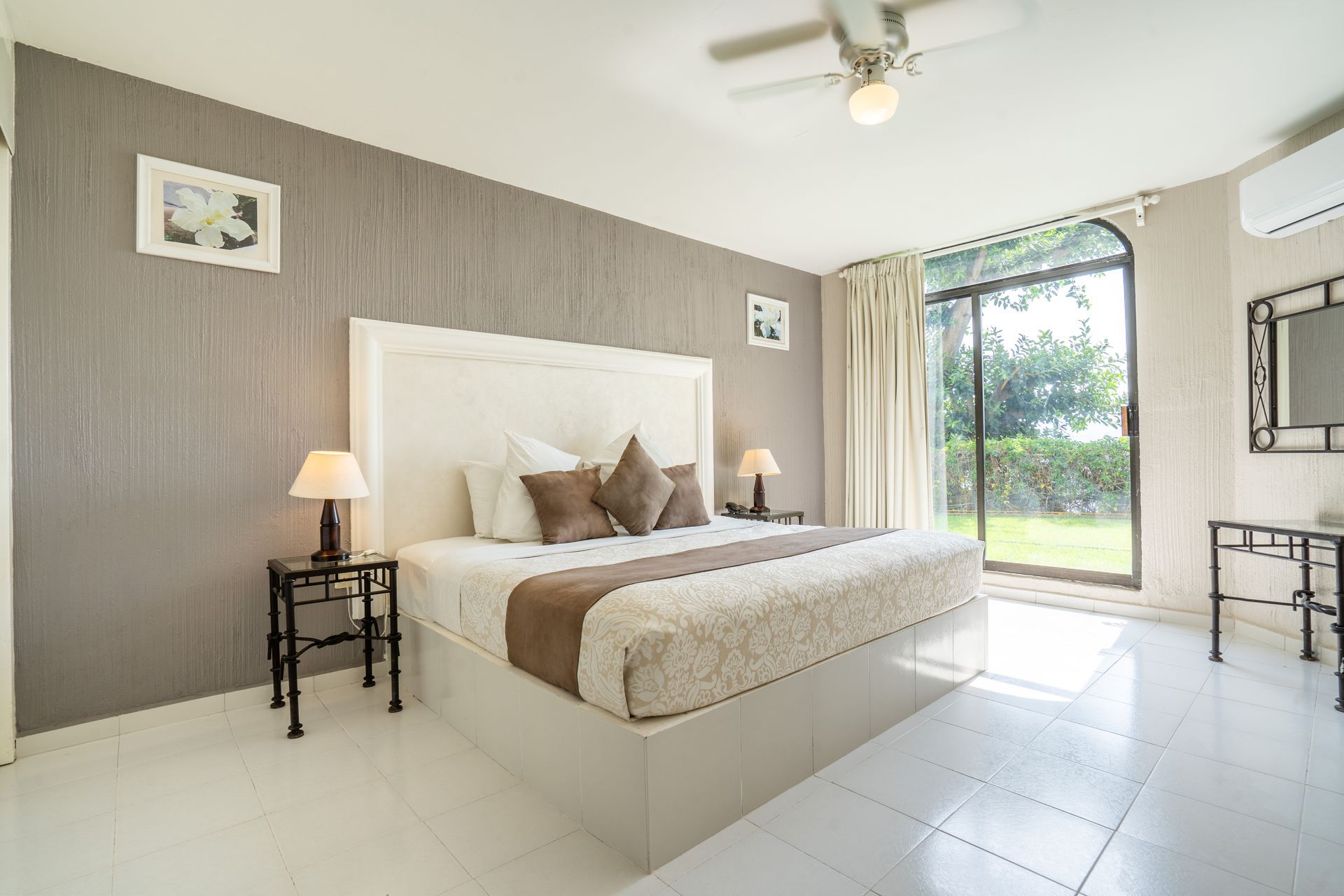 Dormitorio con cama blanca, pared gris y puerta corrediza de vidrio que da a un patio verde.
