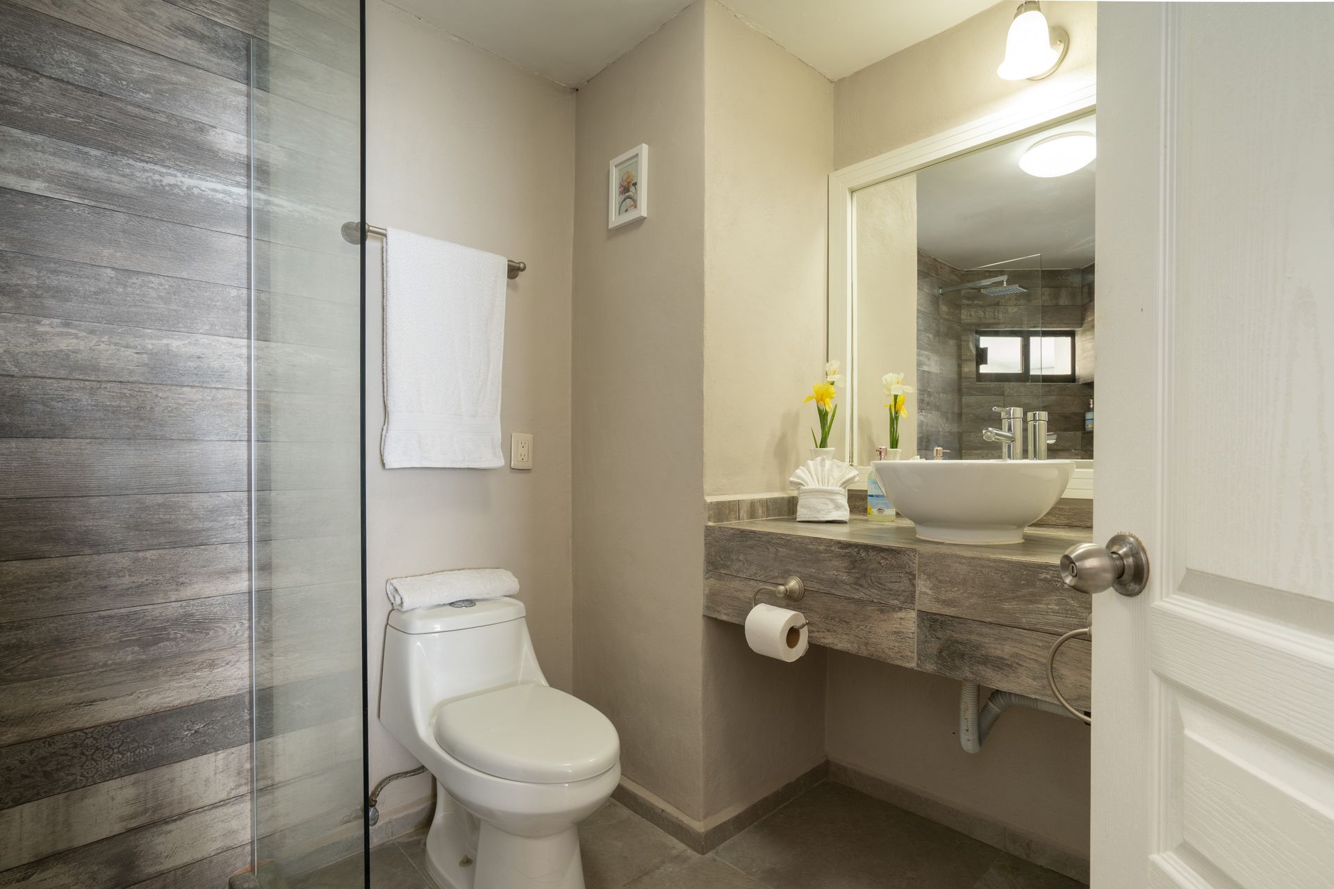 Baño con ducha y tocador con azulejos grises. Inodoro, lavabo y toalla blancos sobre paredes de tonos neutros.
