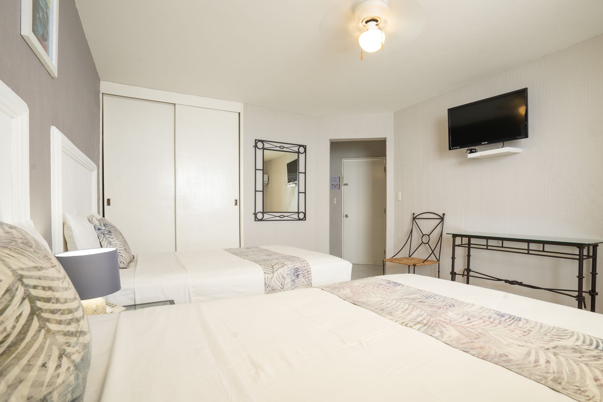 Dos camas en una luminosa habitación de hotel, ropa de cama blanca, televisor, espejo y armario blanco.