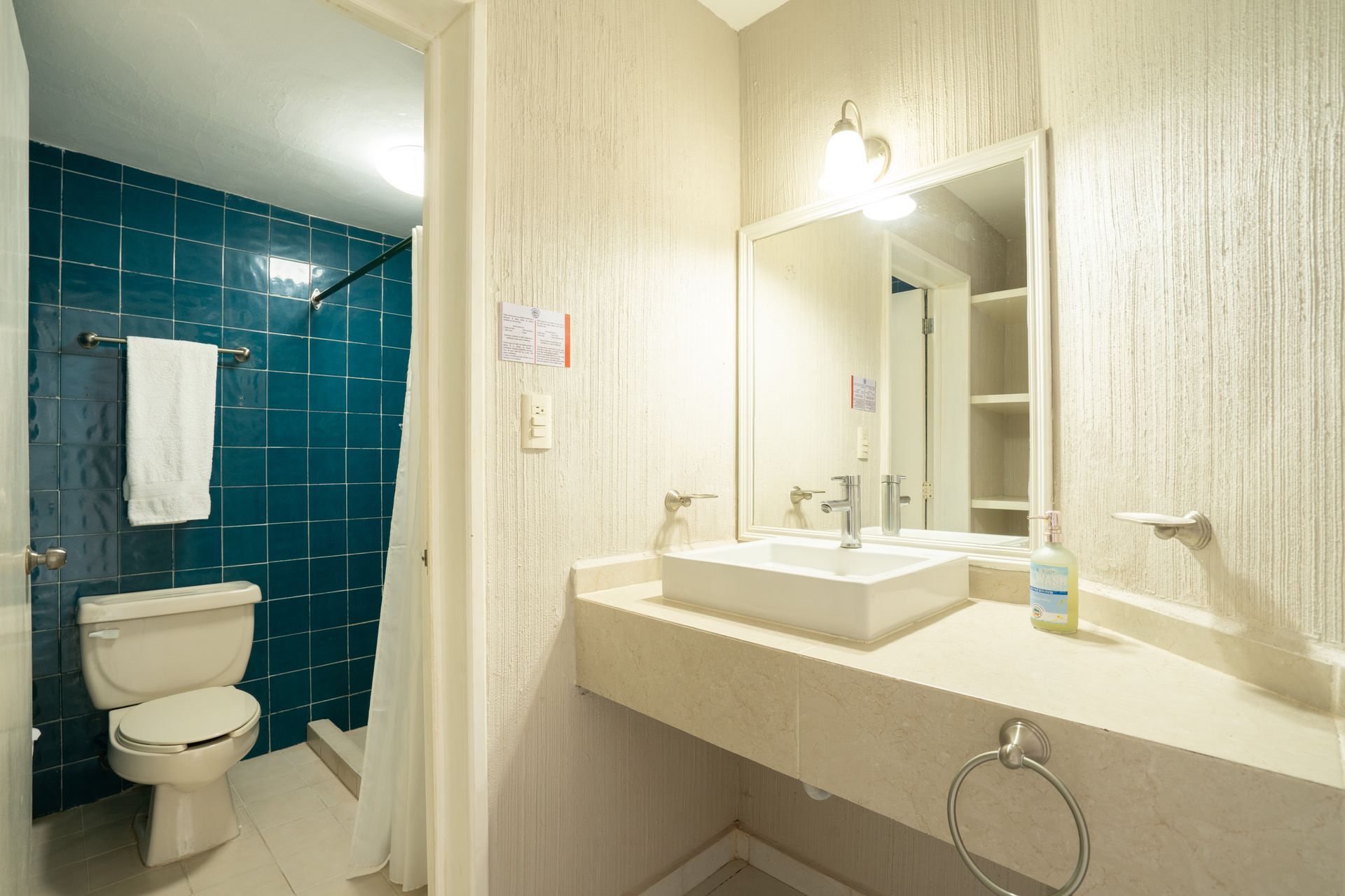 Baño con ducha con azulejos de color verde azulado, inodoro, lavabo, espejo y toallero; paredes neutrales.