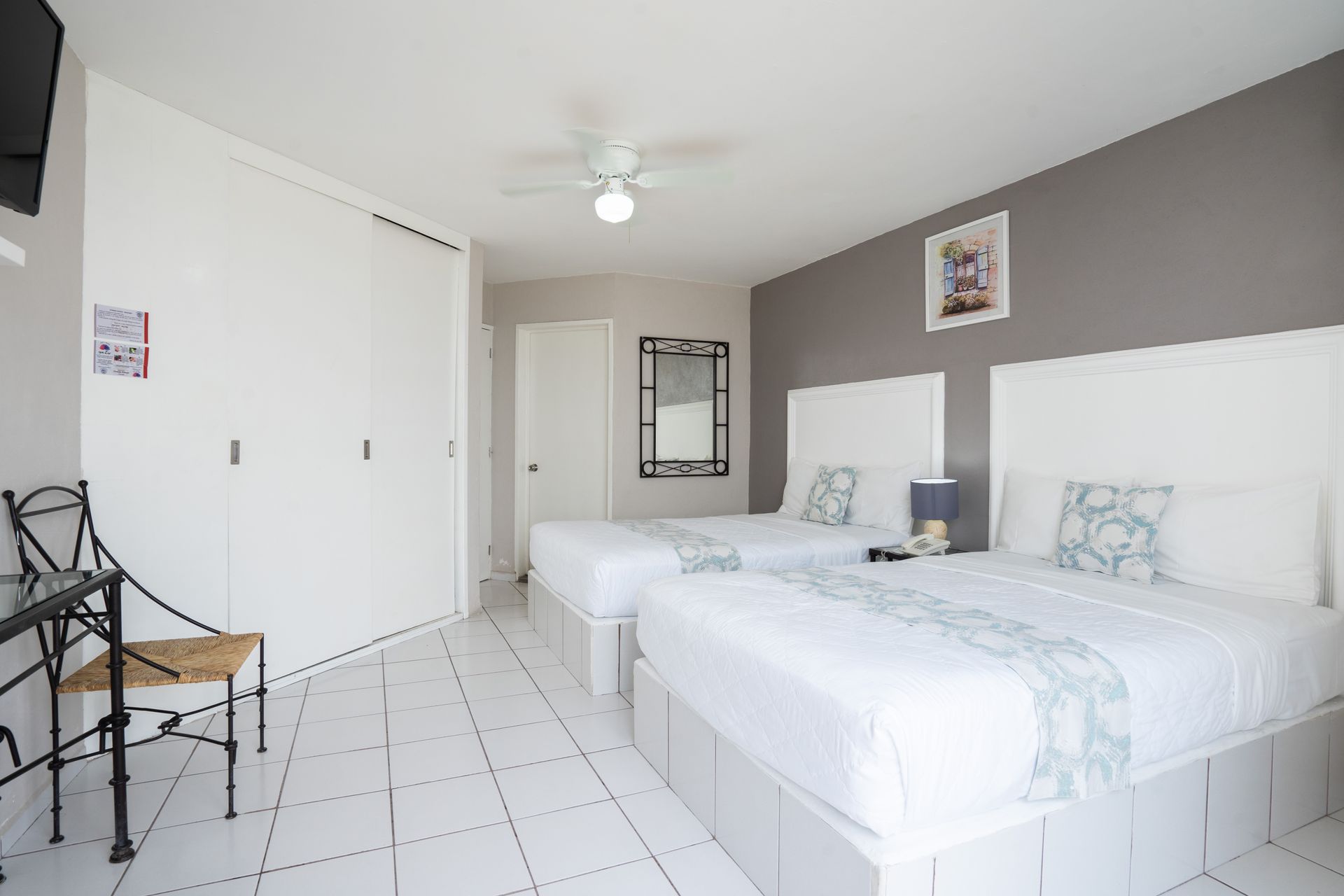 Dormitorio con dos camas, ropa de cama blanca, pared con acento gris, piso de baldosas, armario y mesa pequeña.