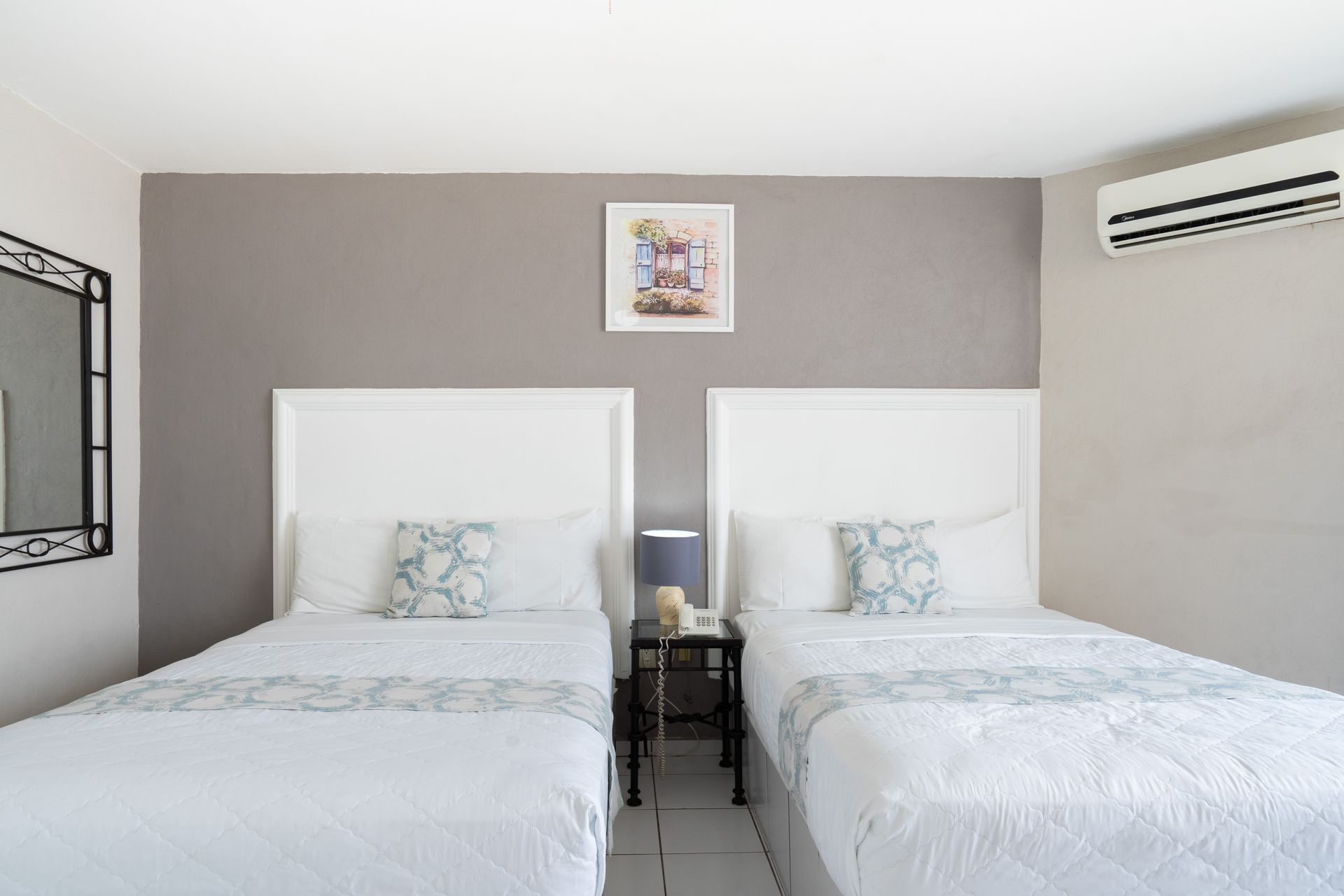 Dos camas individuales en una habitación de hotel, con ropa de cama blanca, pared gris y un pequeño cuadro.