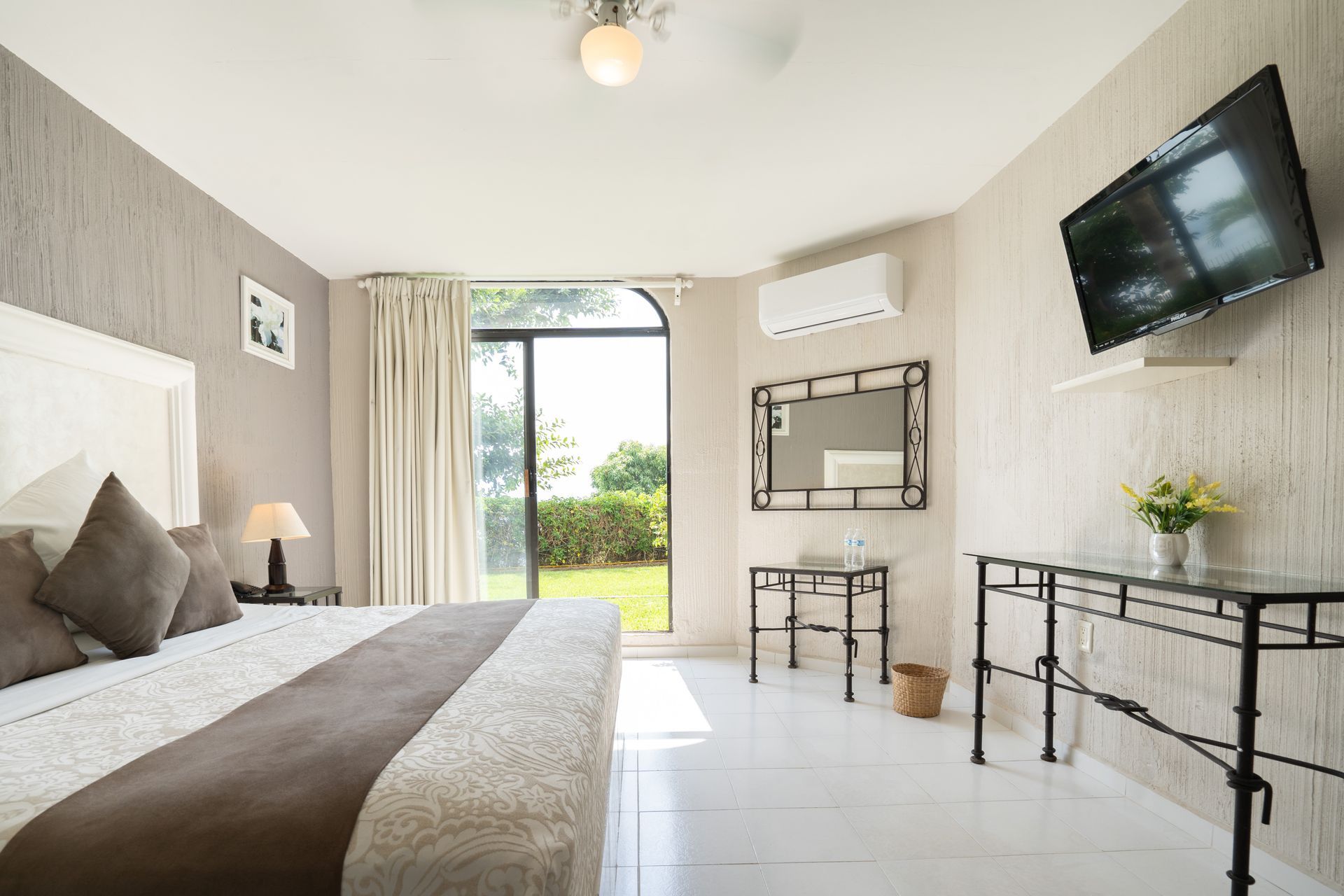 Dormitorio con cama king, televisor, ventana y mesas decorativas. Tonos blanco, marrón y gris.