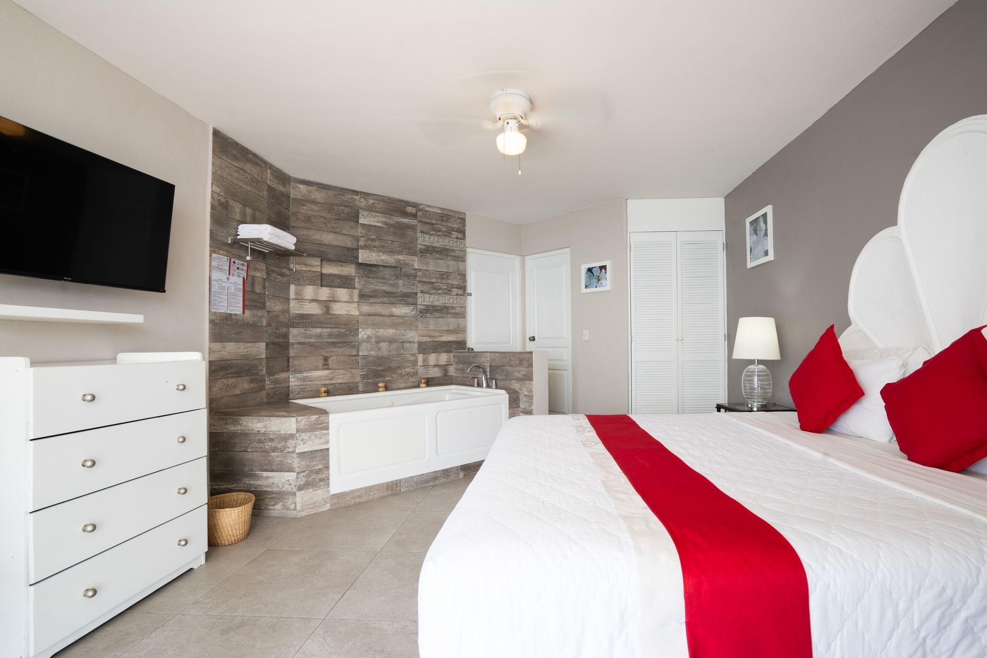 Dormitorio con cama king, bañera, cómoda blanca y televisor. Detalles rojos en las almohadas y el camino de cama.