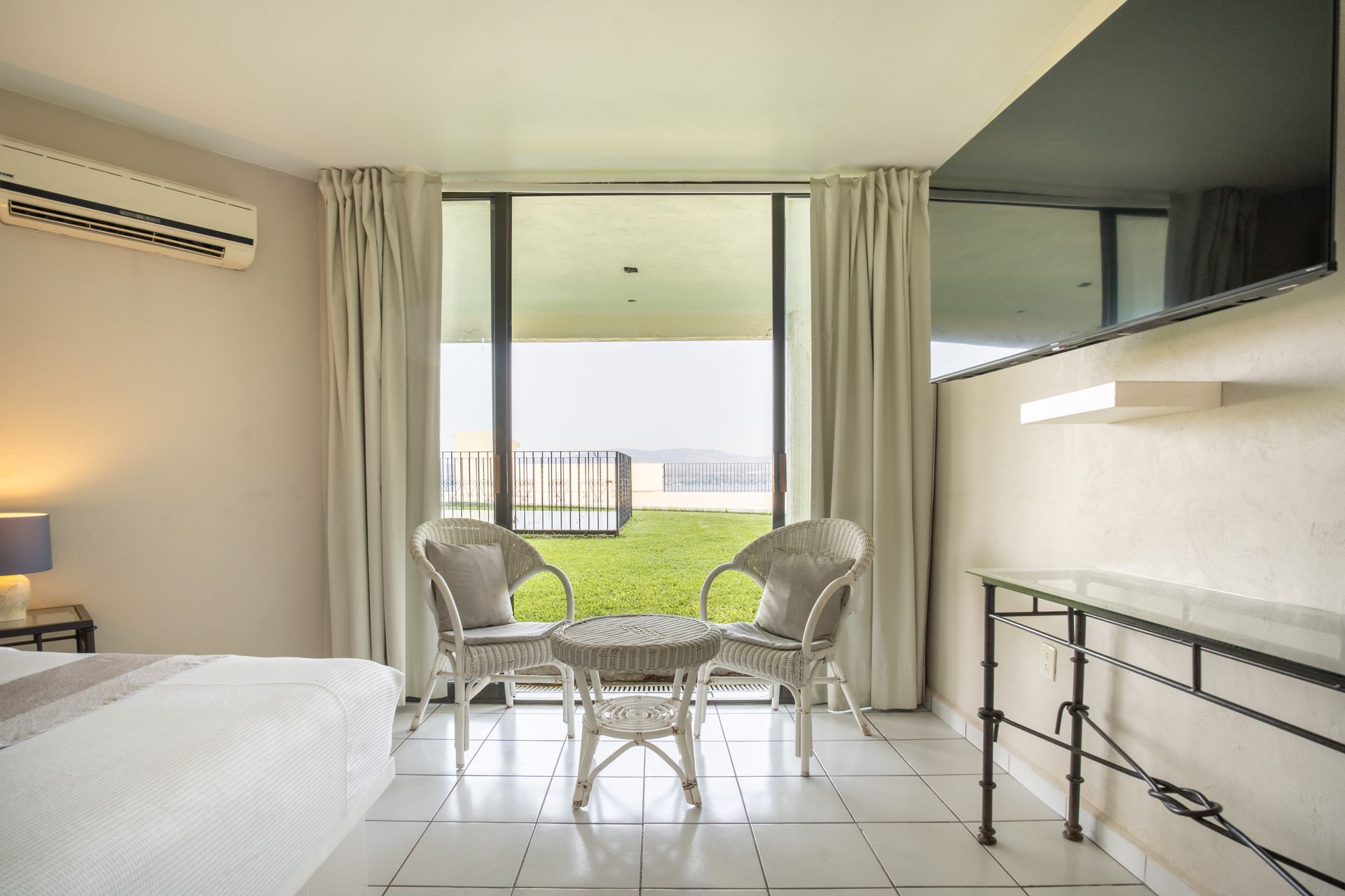 Un dormitorio con sillas de mimbre blancas, mesa y una vista de una zona verde a través de una ventana grande.
