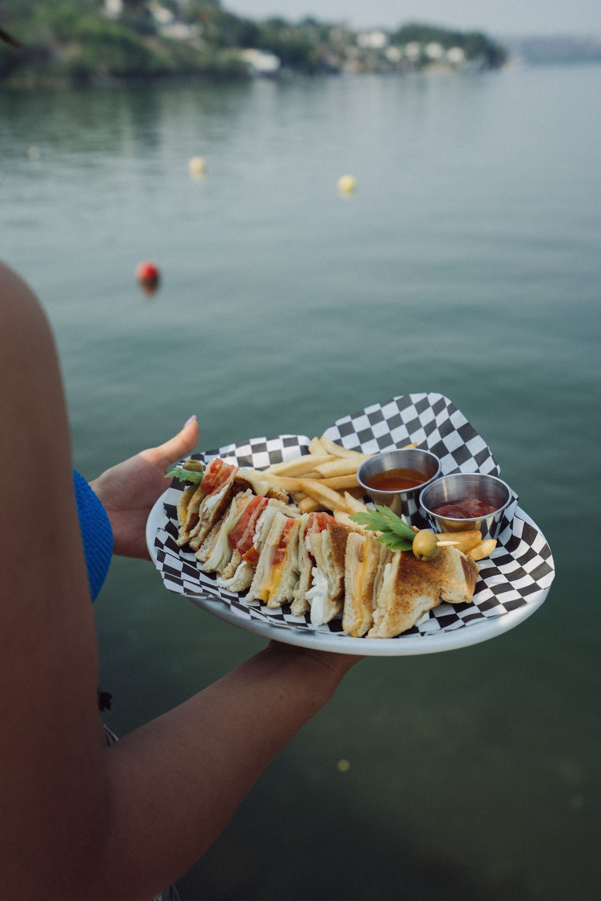Persona que sostiene un plato con un sándwich, papas fritas y salsas para mojar junto a un cuerpo de agua.