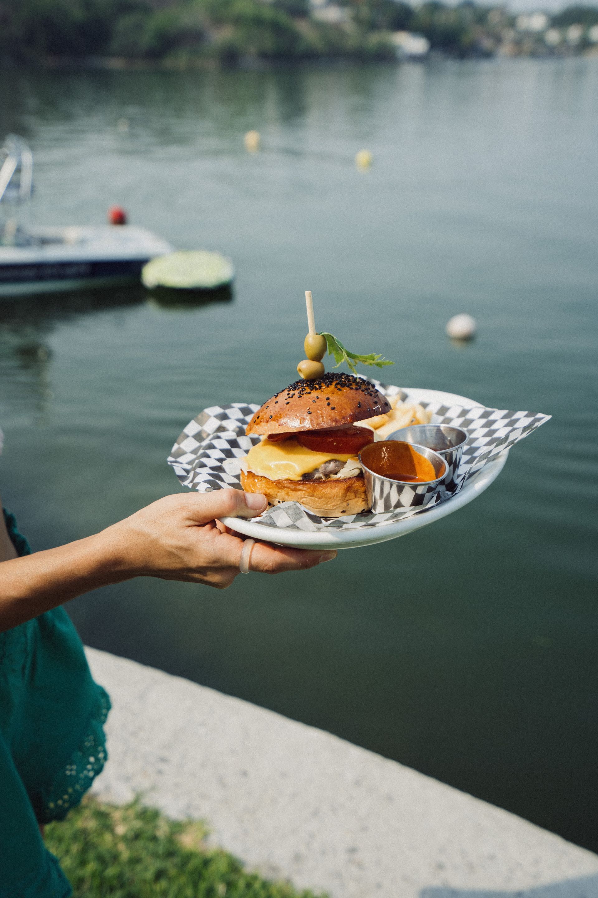 Persona sosteniendo un plato con una hamburguesa y papas fritas junto a un cuerpo de agua.