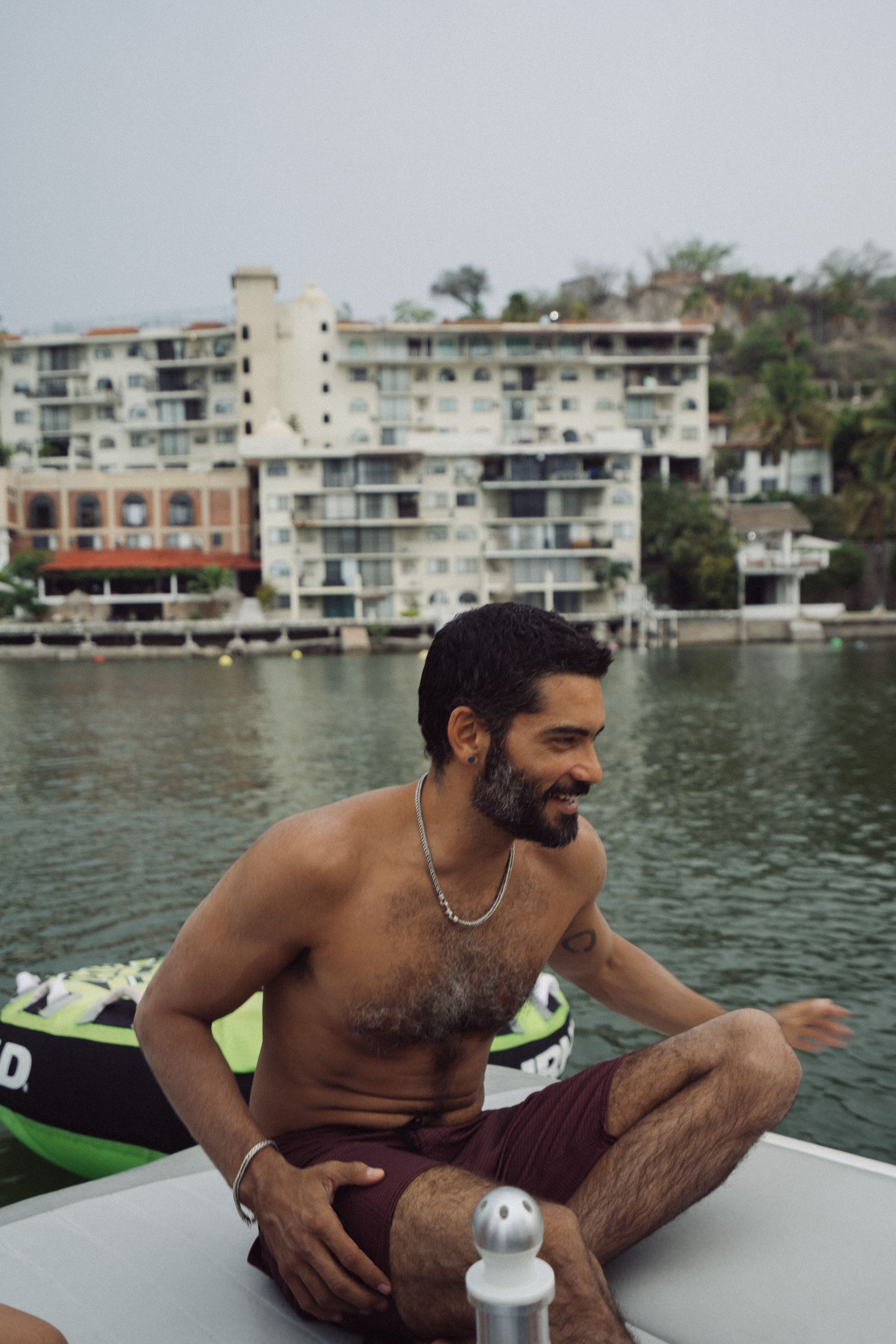 Hombre de pelo oscuro y barba en un barco, sonriendo, sin camisa y con pantalones cortos. Edificios y agua al fondo.