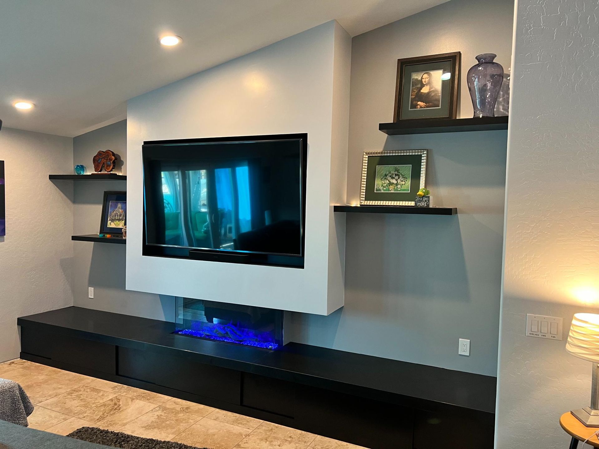 Built-in entertainment center with a floating TV, black cabinets, shelves with decor, and a blue fireplace glow.