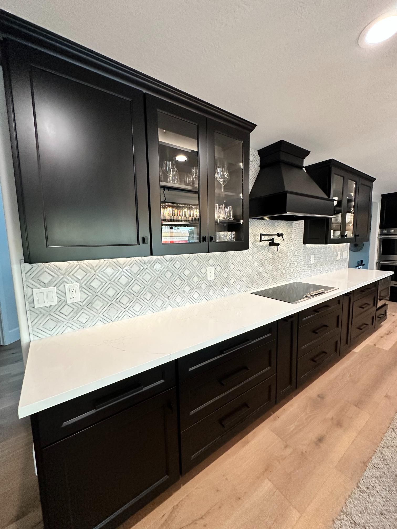 Black kitchen cabinets with white countertops, backsplash, and wooden floors.
