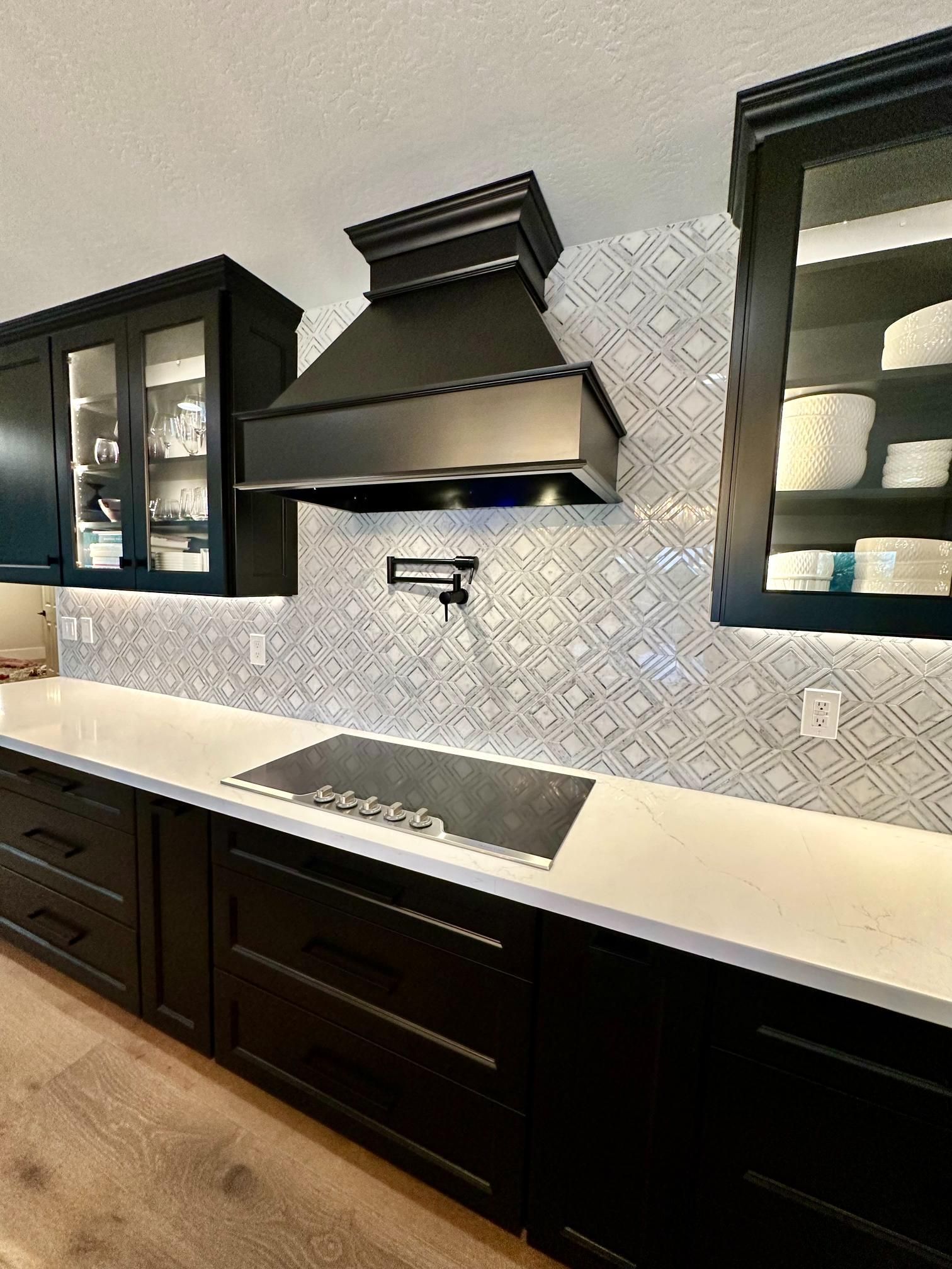 Black kitchen with a range hood, white countertop, black cabinets, and patterned backsplash.