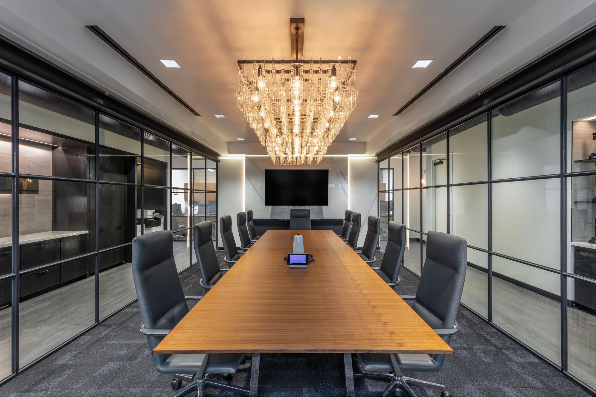 Long conference room with a wooden table, black chairs, and a large chandelier. Glass walls and a black screen.
