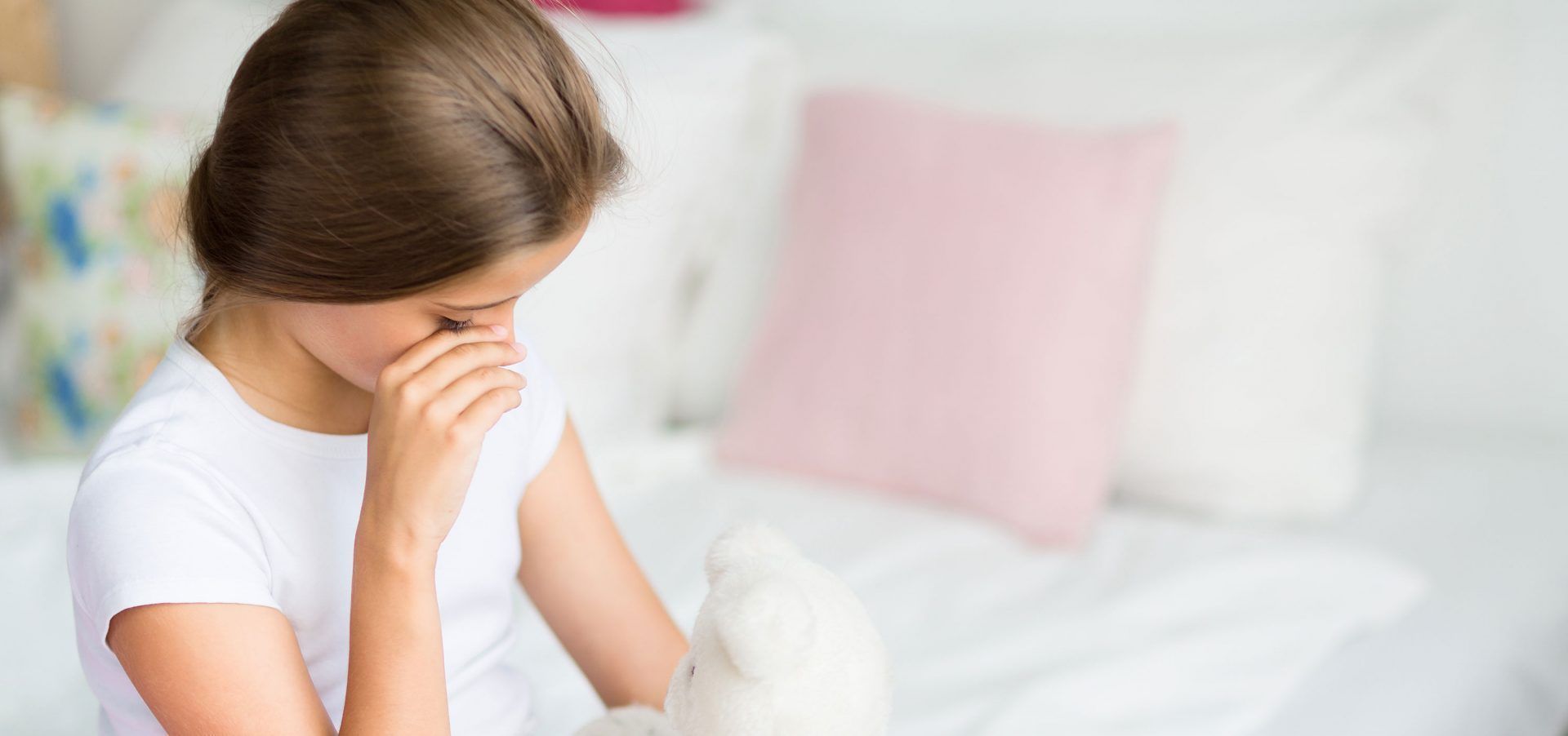 Person sitting on a bed, covering their face with hands, appearing upset in a soft pink bedroom.