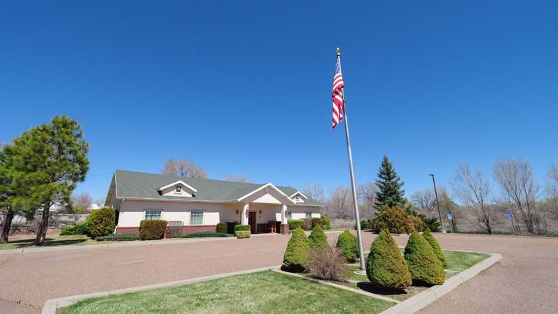 Single-story building with an American flag, landscaped shrubs, and a driveway under a clear blue sky